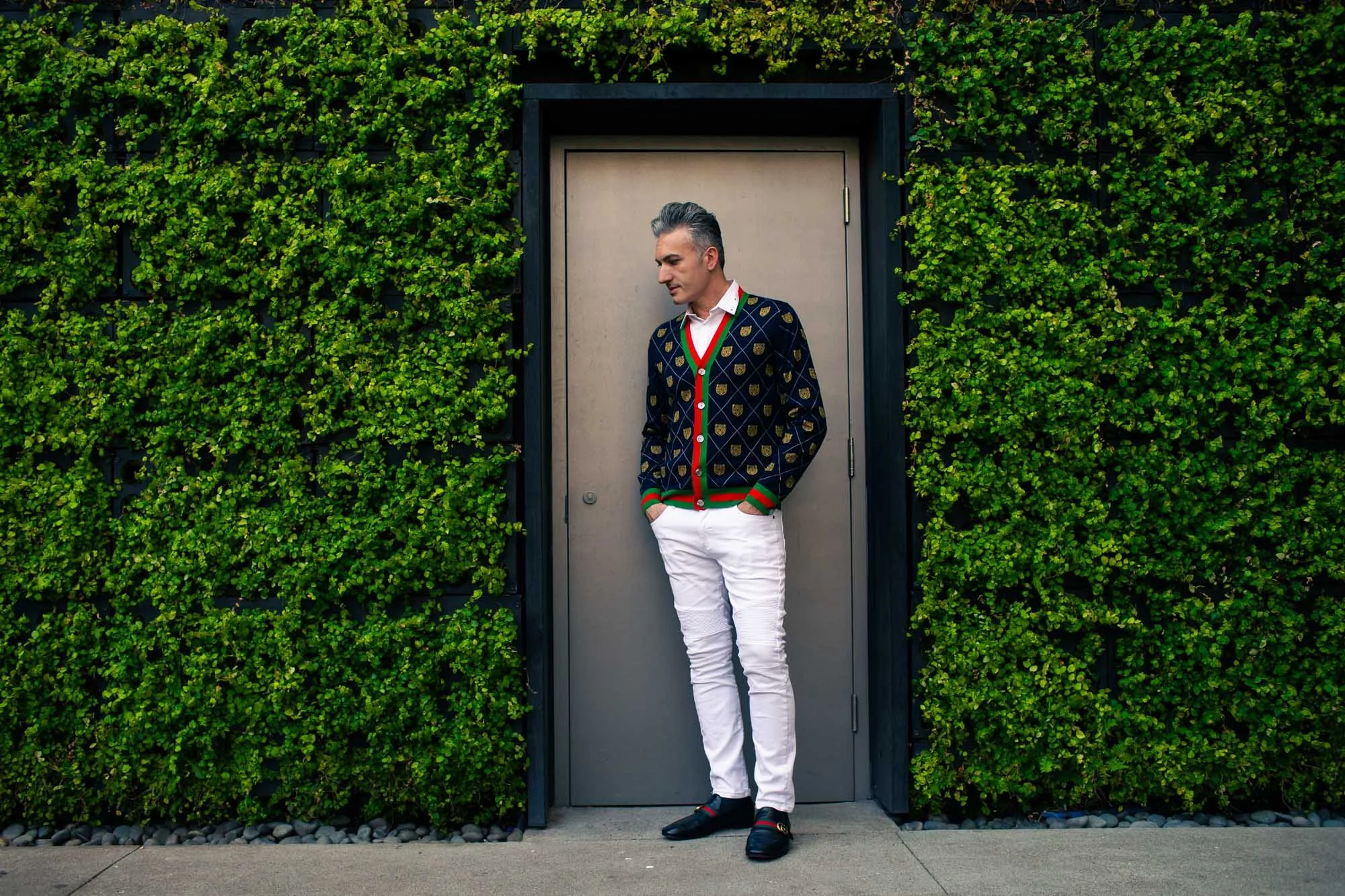 A man standing against a gray door surrounded by vibrant green foliage. He is wearing a navy blue jacket with gold patterns, a white shirt, white pants, and black shoes with red and green accents.
