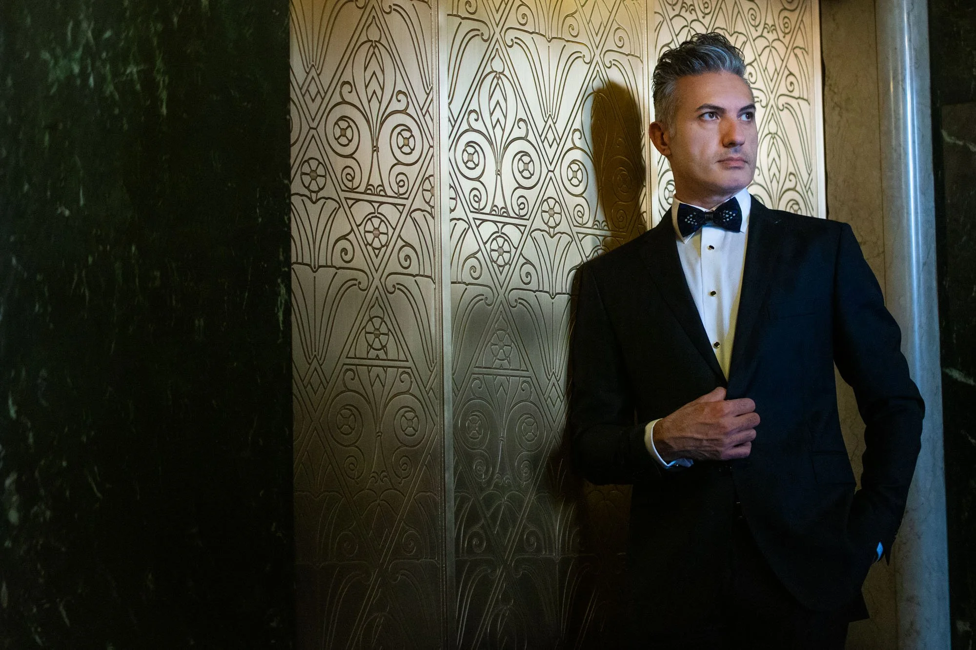 A man in a tuxedo with a bow tie standing against a decorative gold wall in an elevator or similar setting.