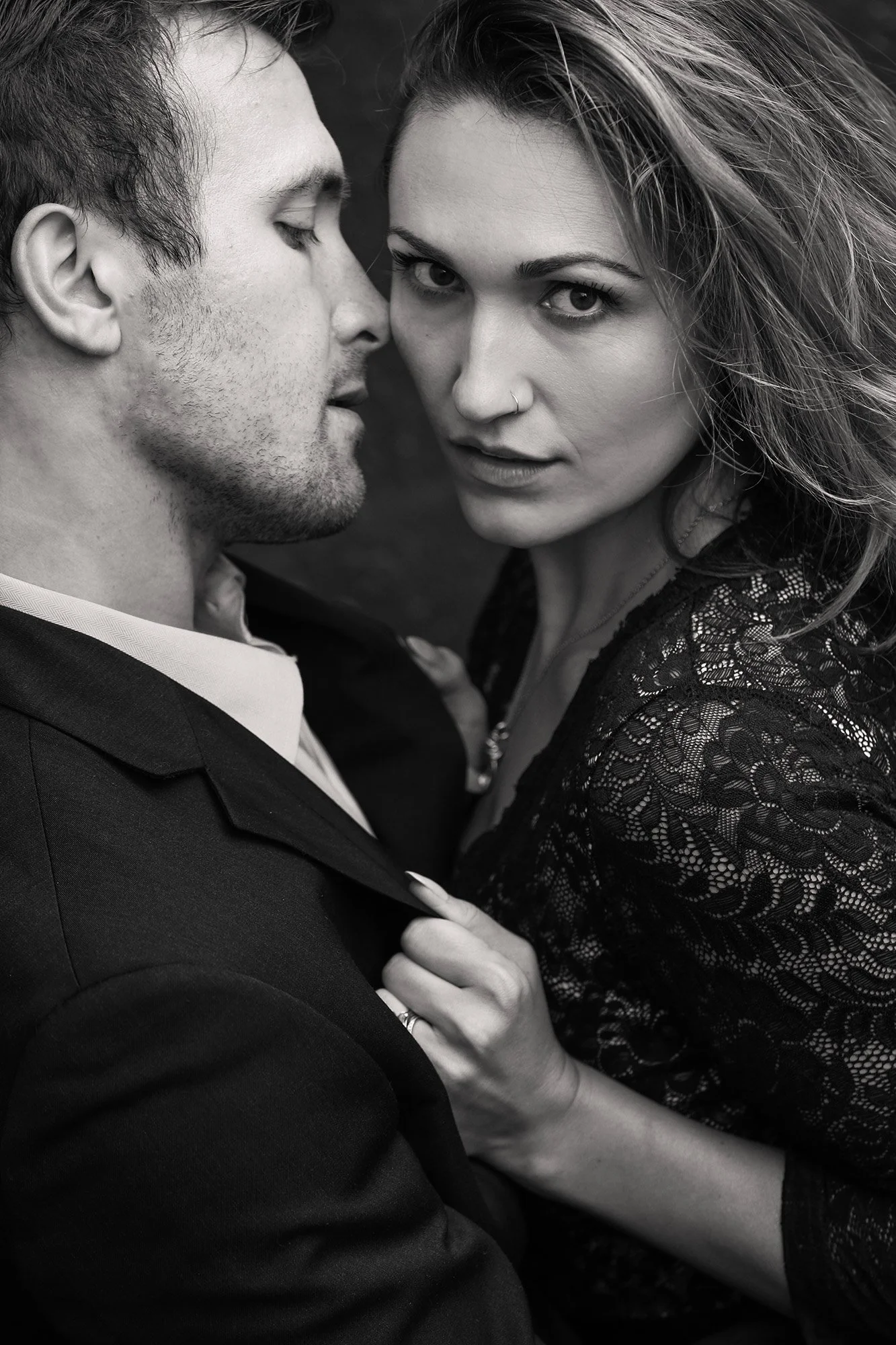 A black-and-white photo of a man and woman in an intimate pose. The man is on the left, his eyes closed, slightly smiling, with short, curly hair and a beard. The woman is on the right, looking directly at the camera with a confident expression, hold