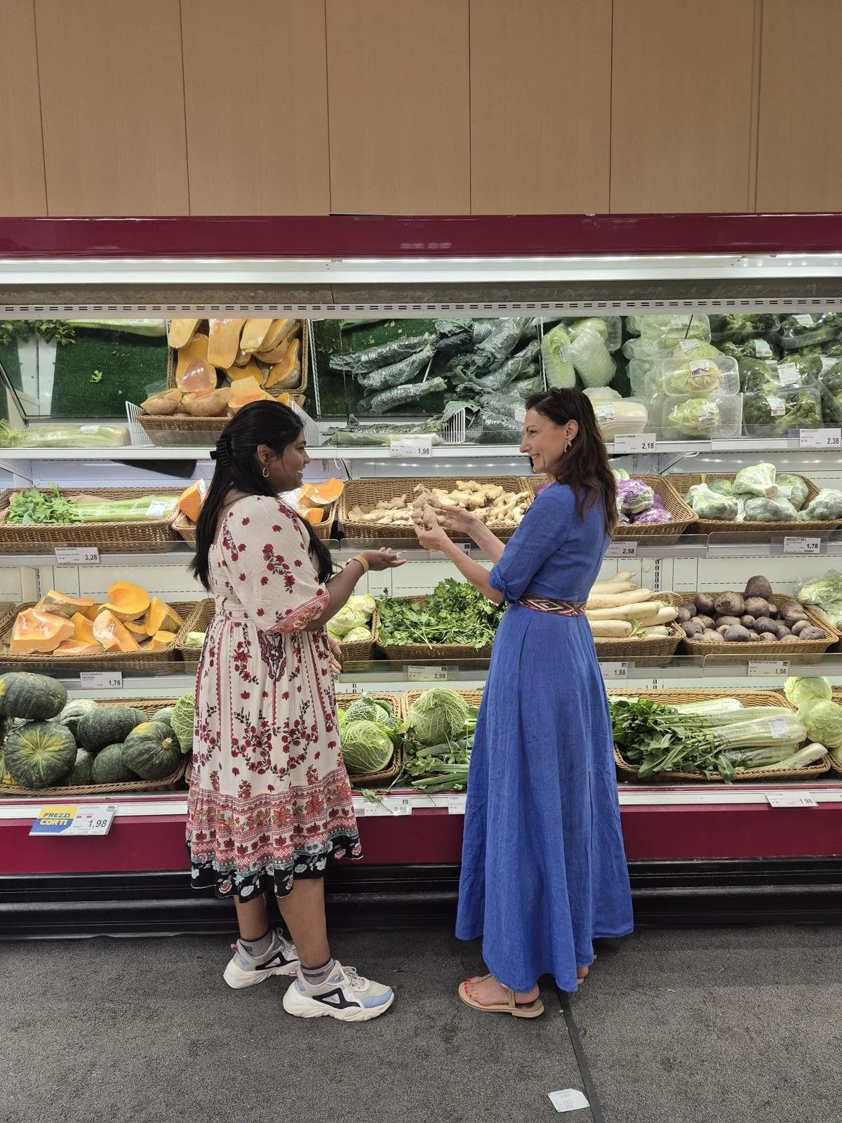 Two women shopping for vegetables in a grocery store, with produce like squash, zucchini, ginger, beets, and cauliflower displayed behind them.