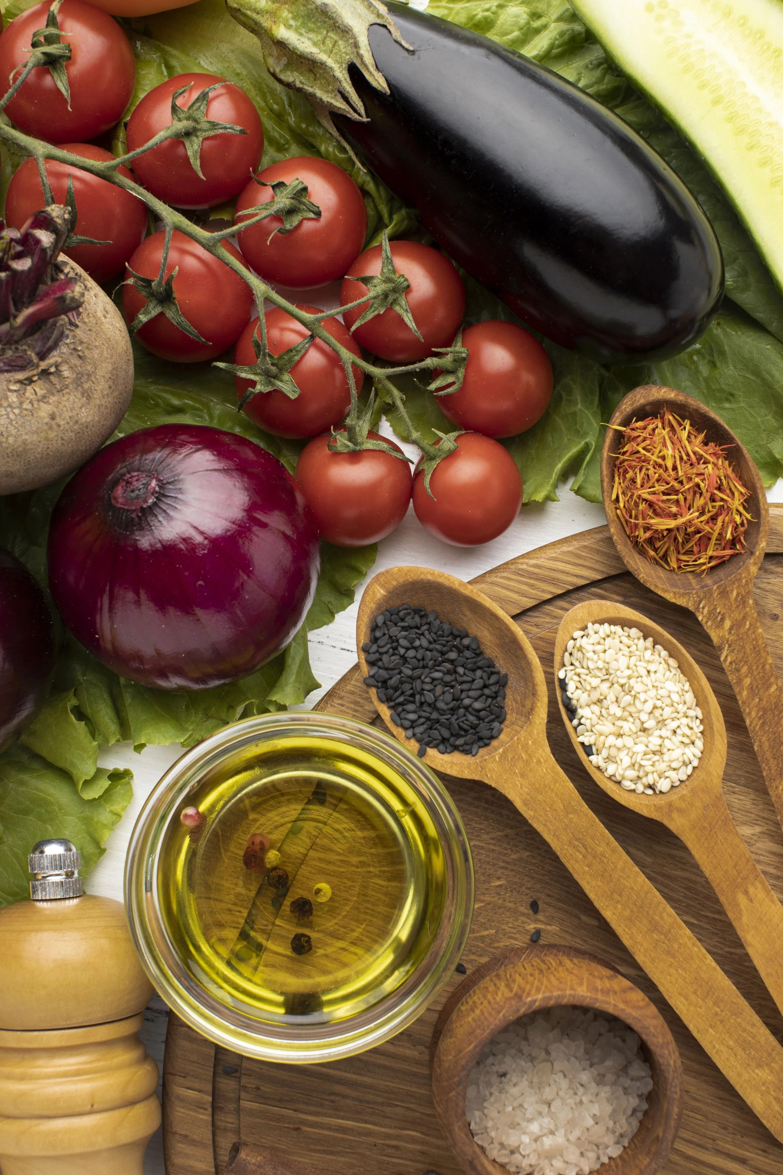 Fresh vegetables and spices arranged on a wooden surface, including cherry tomatoes, eggplant, cucumber, red onion, lettuce, and various seeds, oils, and seasonings in small wooden bowls and spoons.
