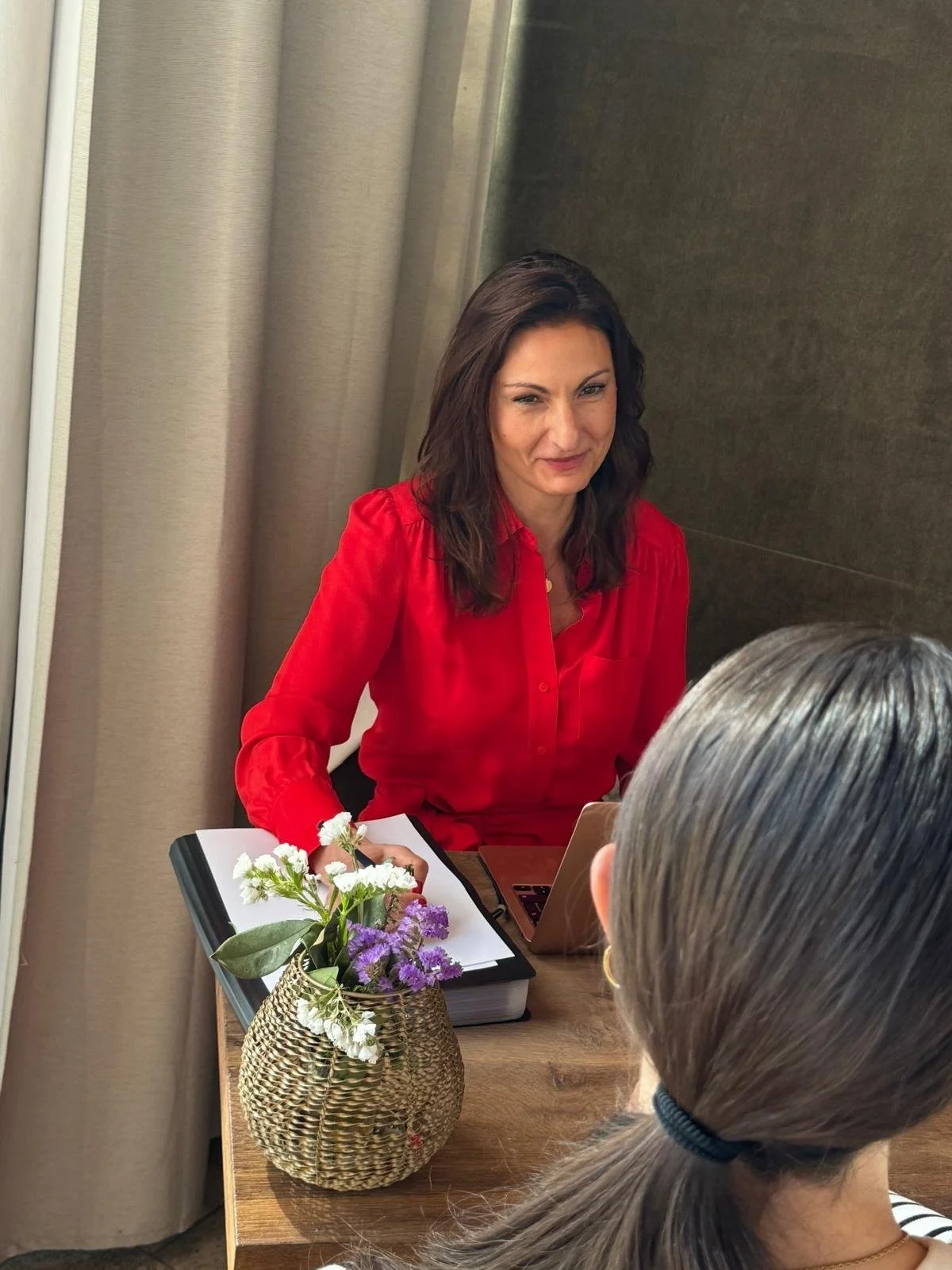 A woman with dark hair and a red blouse sitting at a desk, talking to another woman with gray hair. There is a notebook, a laptop, and a vase with flowers on the desk.