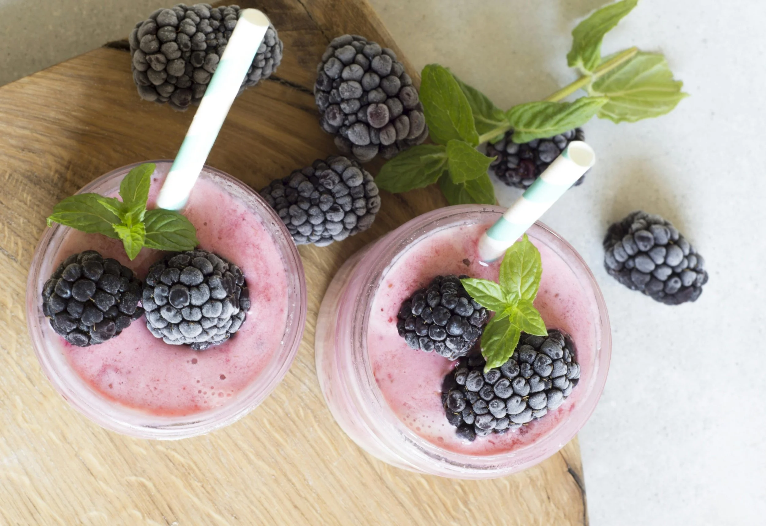 Two glasses of blackberry smoothies garnished with fresh blackberries and mint leaves, with striped straws, placed on a wooden tray with additional blackberries and a sprig of mint nearby.
