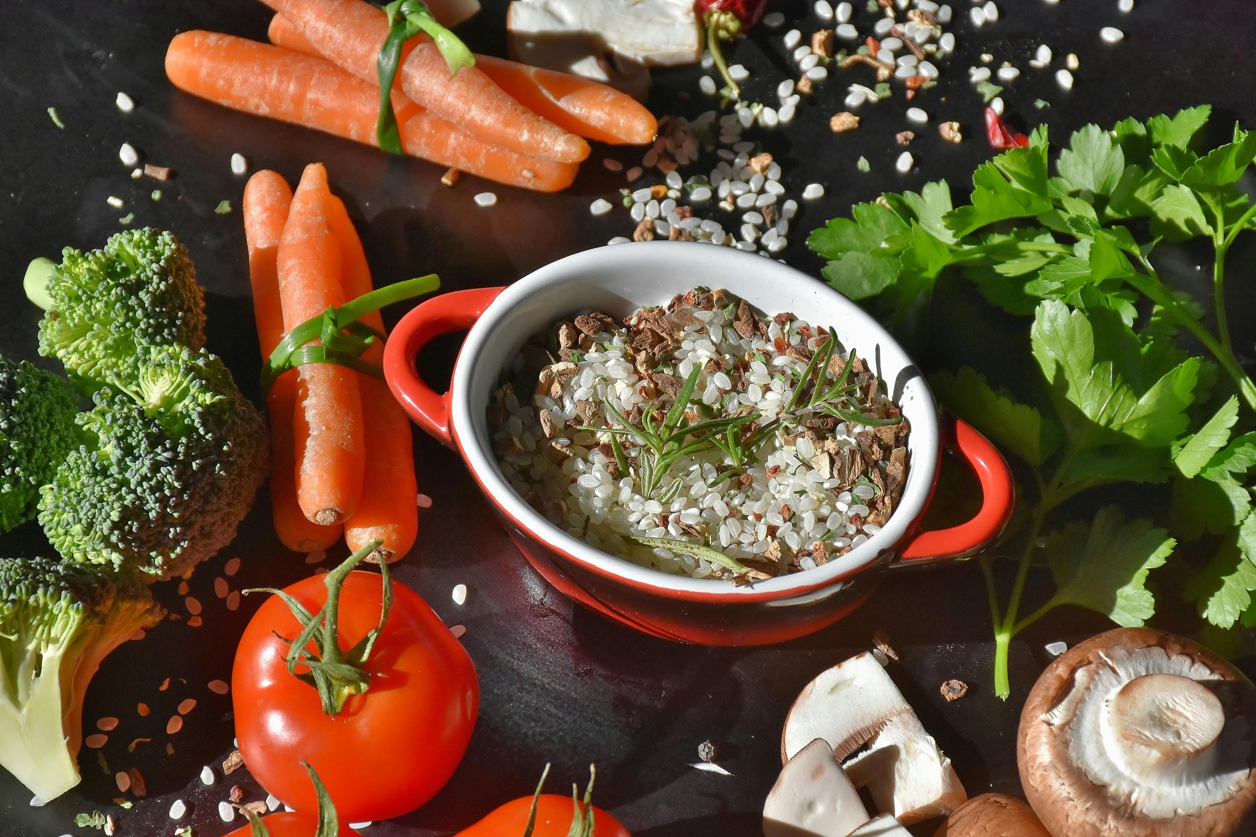 Fresh vegetables including cherry tomatoes, broccoli, carrots, and mushrooms surrounding a bowl of sprouted lentils or grains on a black surface.