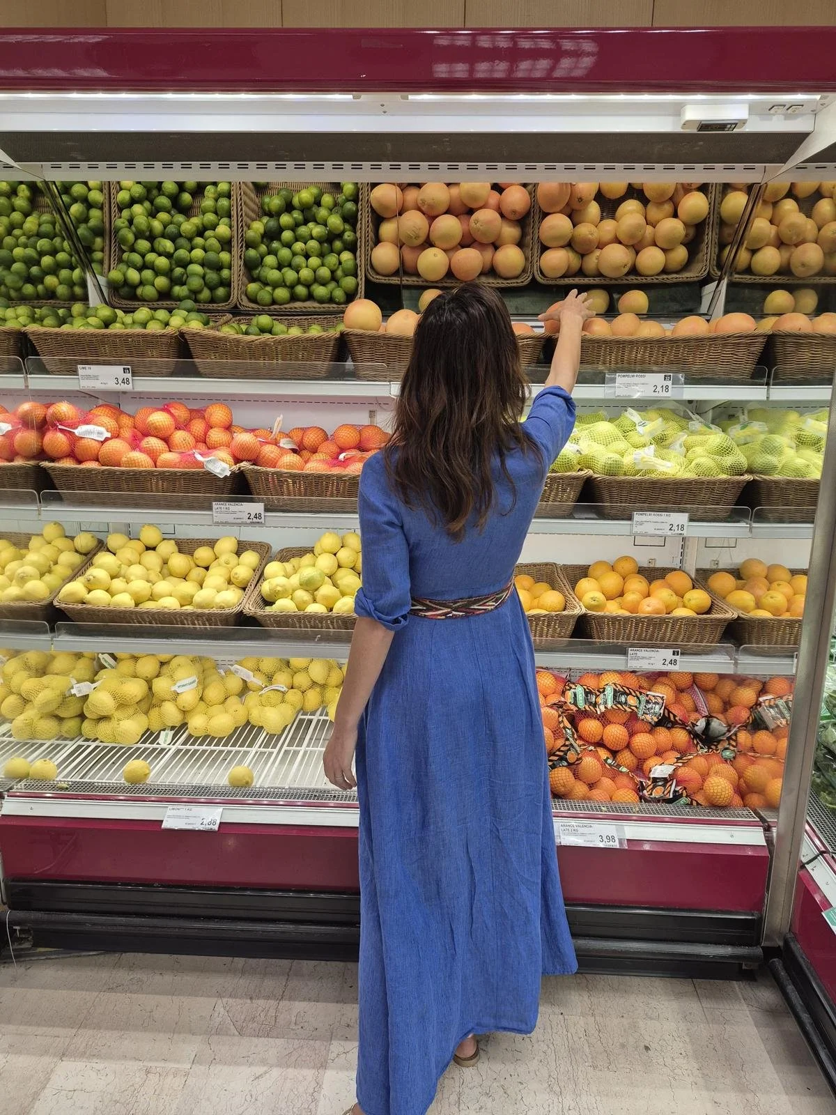 A woman in a blue dress shopping for fruit in a grocery store, reaching for a peach from the produce section.