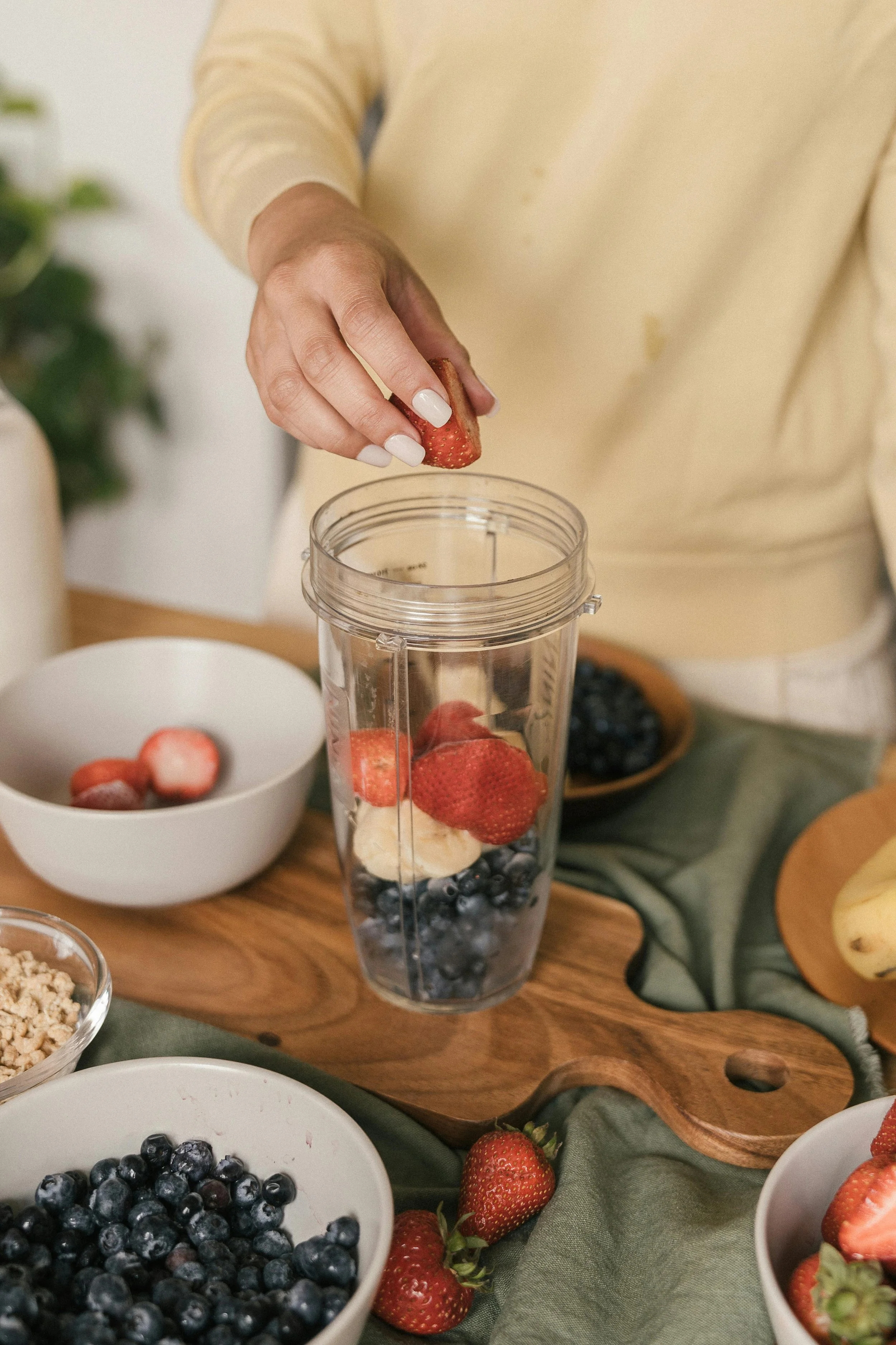 Person placing strawberries into a blender with blueberries, banana slices, and other berries on a wooden cutting board, surrounded by bowls of fruit.