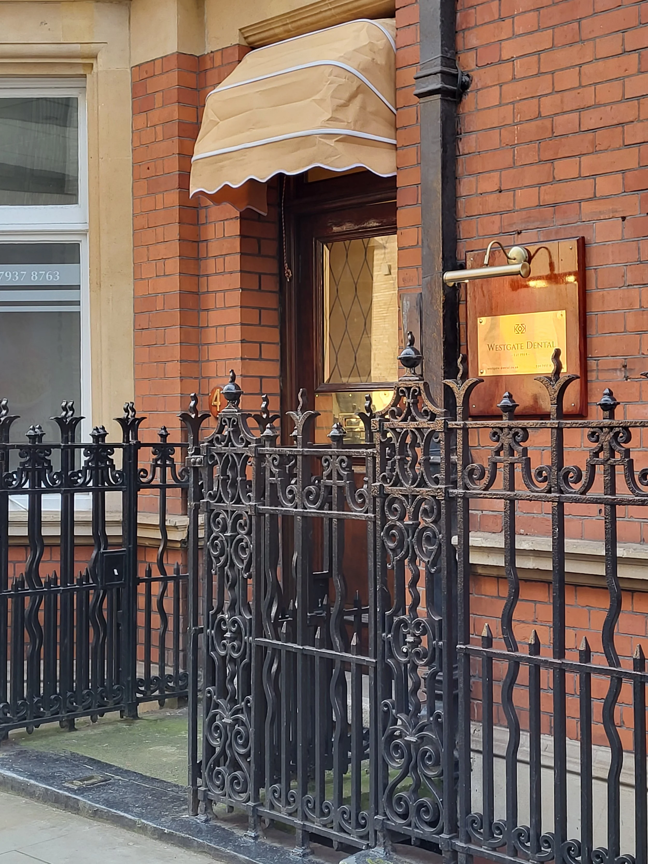The entrance to Westgate Dental features a brick building with a decorative black wrought iron fence in front. There is a window on the left with horizontal blinds, and a beige and white striped awning above the door. A brass-colored sign reads 'Westgate Dental' and is mounted on a wooden board to the right of the door.