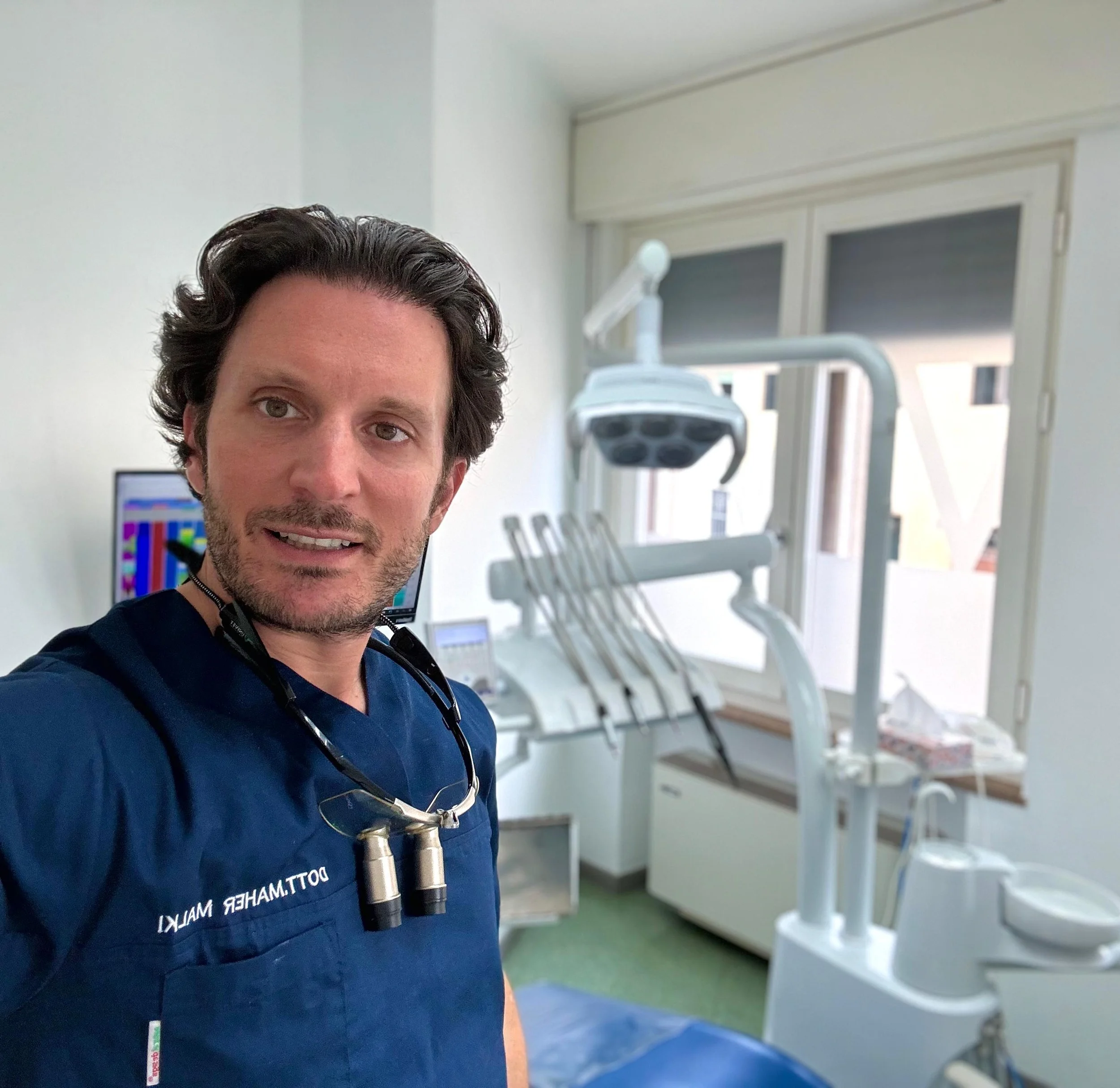 A male dentist in scrubs with a pair of dental loupes around his neck standing in a dental clinic with dental equipment and a window in the background.
