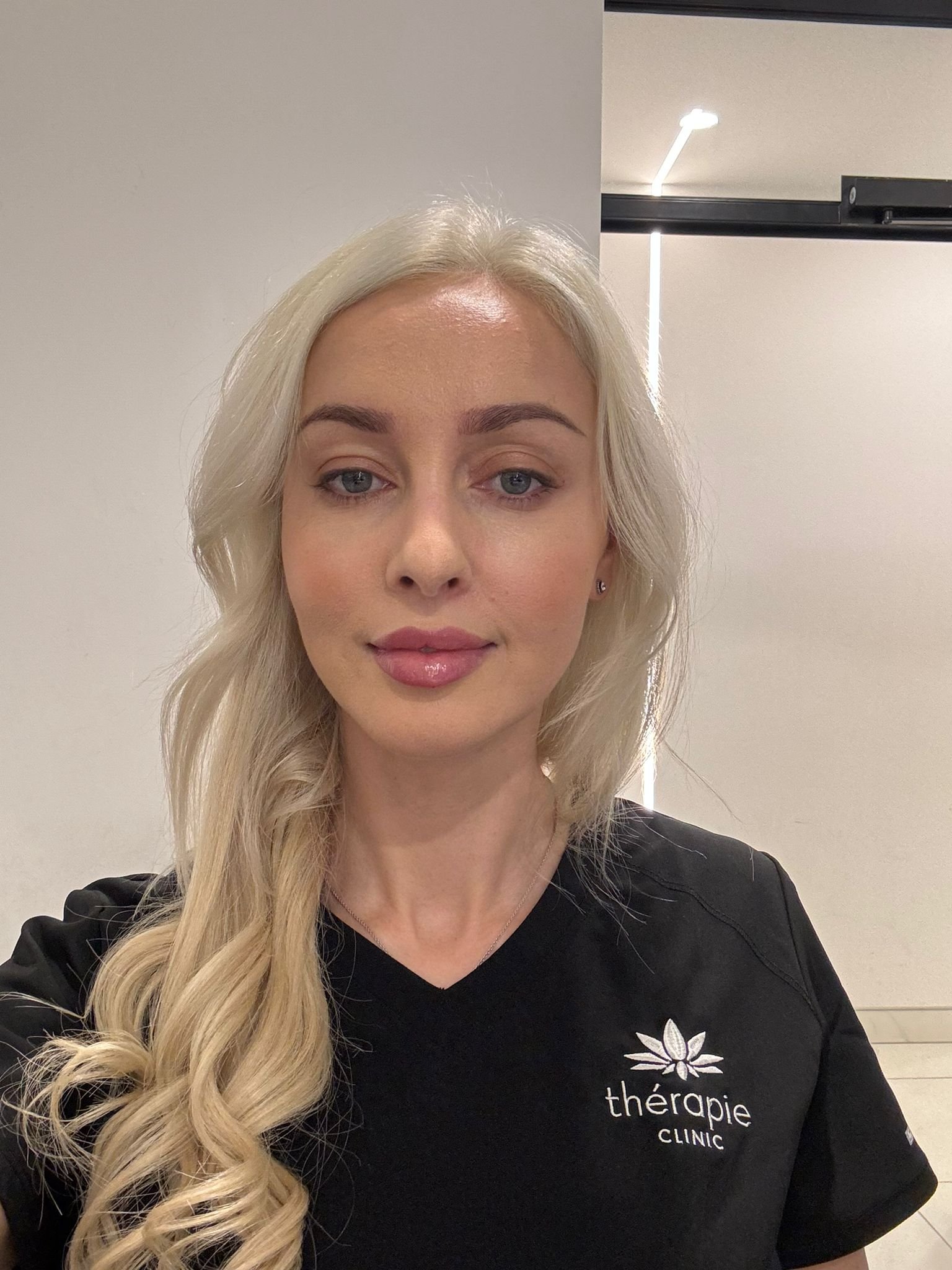 A woman with long, wavy blonde hair wearing a black scrub top standing in front of a plain wall and partially visible door.