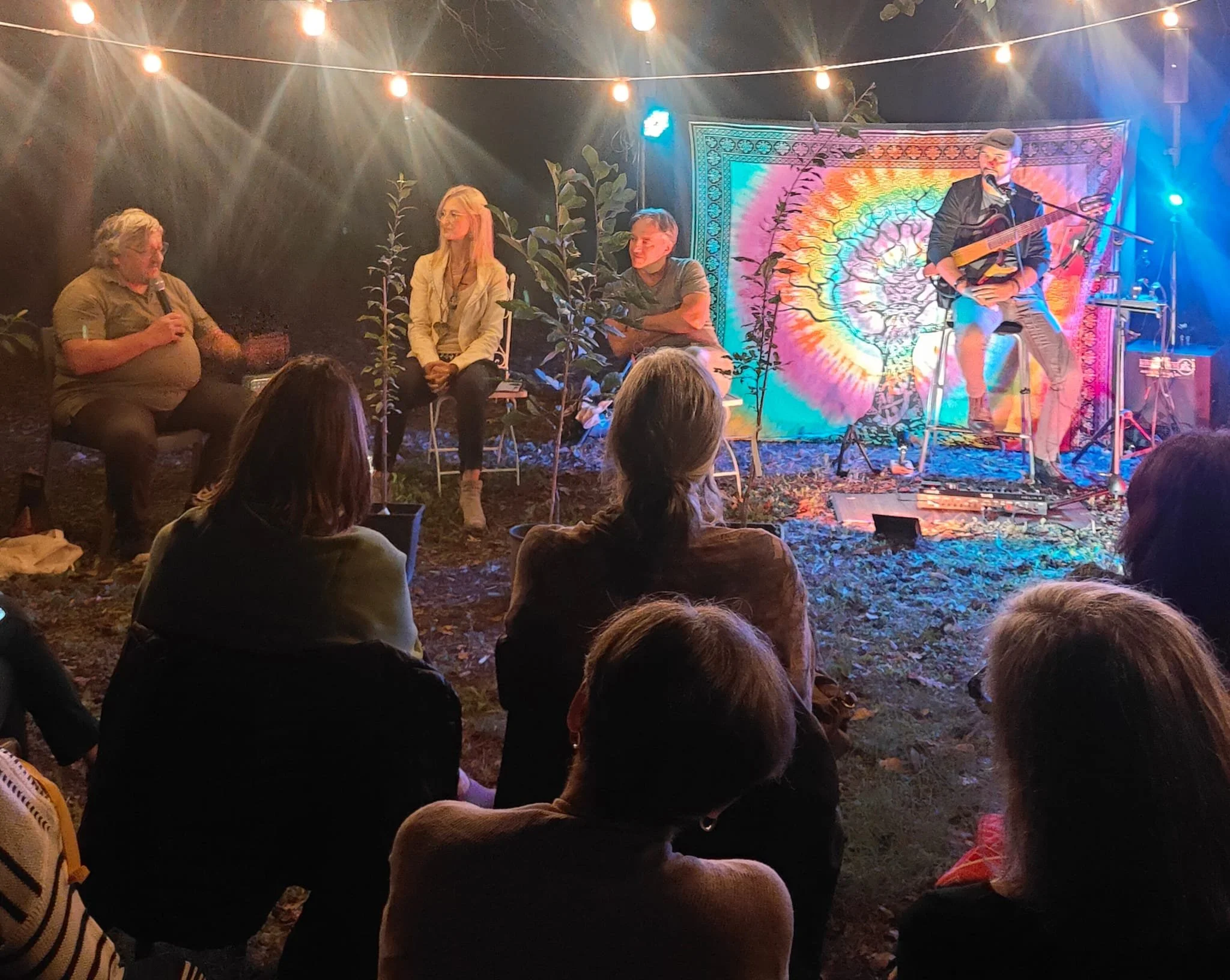 Un gruppo di persone ascolta un concerto in un ambiente all'aperto con luci calde, decorazioni colorate e piante. C'è un musicista con chitarra acustica che suona e altre persone sedute a discutere.
