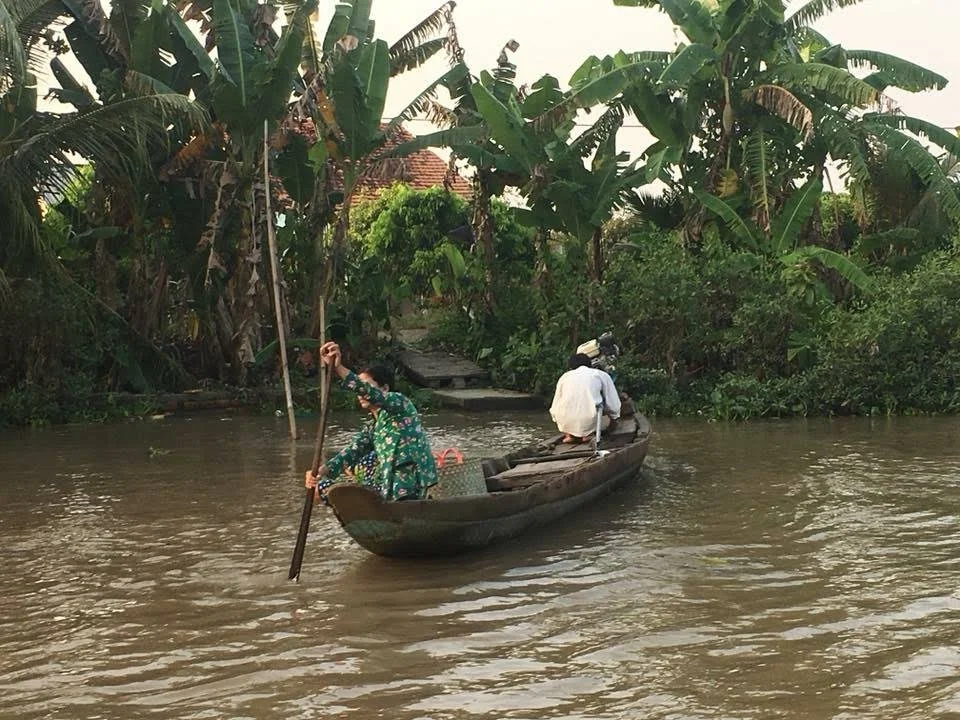 Deux personnes en bateau sur une rivière entourée de végétation tropicale, avec des bananiers. L'une des personnes porte un masque et une robe à motifs floraux, tandis que l'autre est en blanc avec un chapeau.