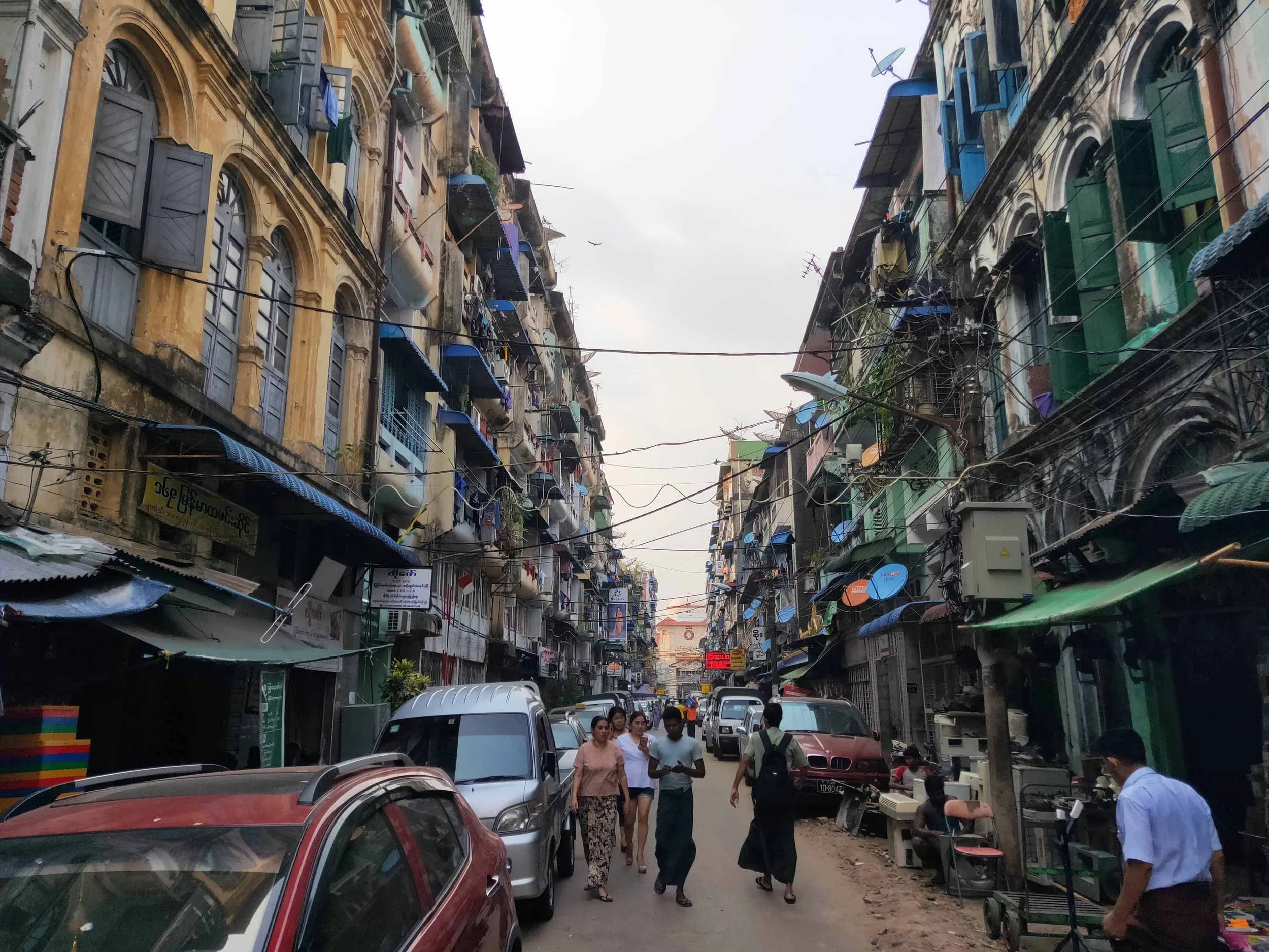 Rue urbaine étroite bordée d'immeubles anciens avec fenêtres à volets colorés et appareils de climatisation, voitures stationnées, et des passants marchant dans la rue, sous un ciel nuageux.