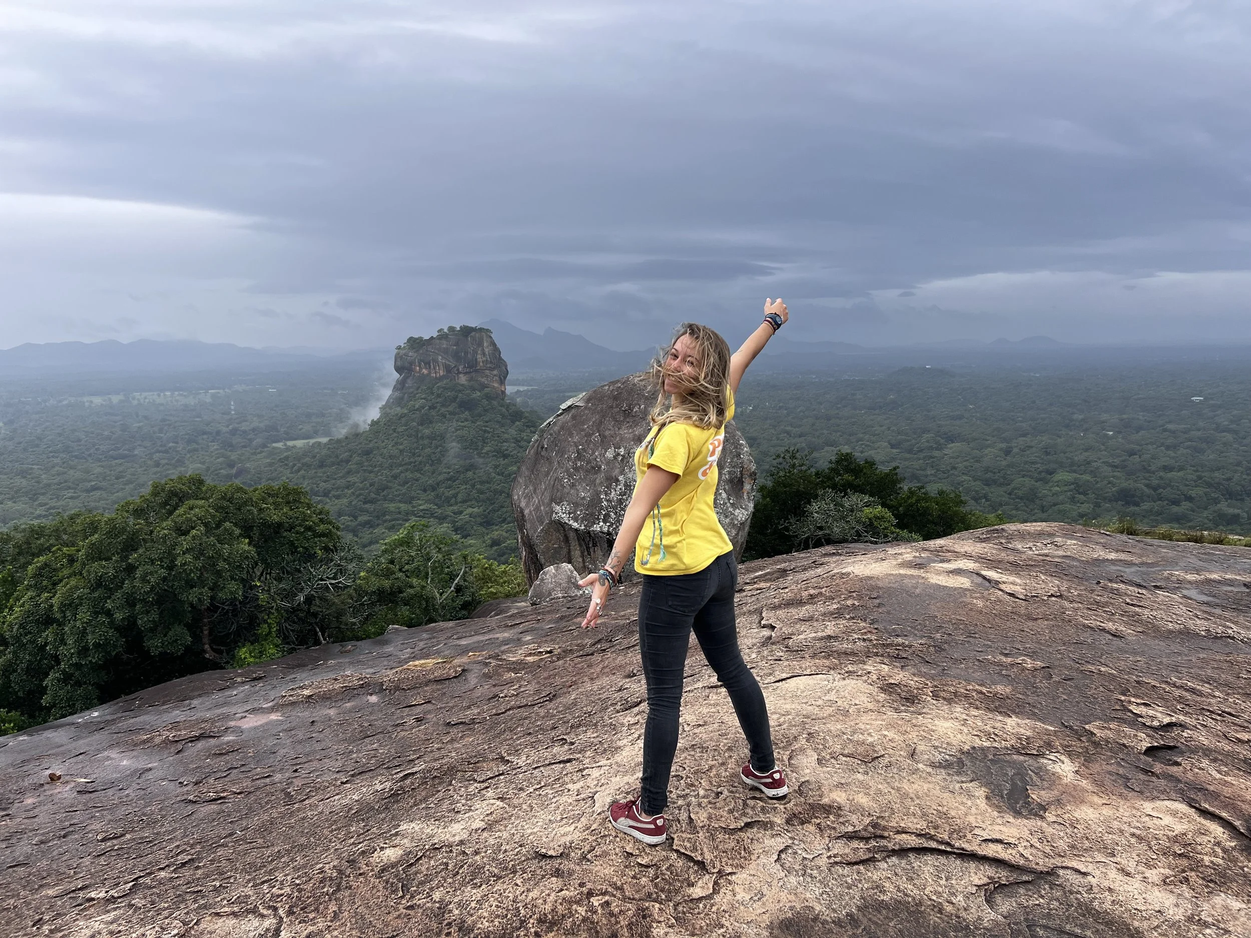Une femme souriante en t-shirt jaune et pantalon noir pose sur un rocher en montagne, avec un paysage de forêt dense et une formation rocheuse spectaculaire à l'arrière-plan, sous un ciel nuageux.