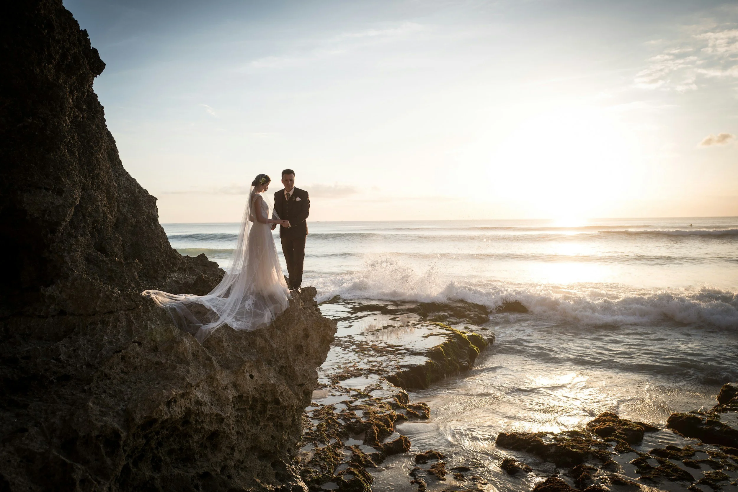 Un couple de mariés, une femme en robe blanche et un homme en costume noir, se tenant la main sur un rocher au bord de la mer au coucher du soleil.