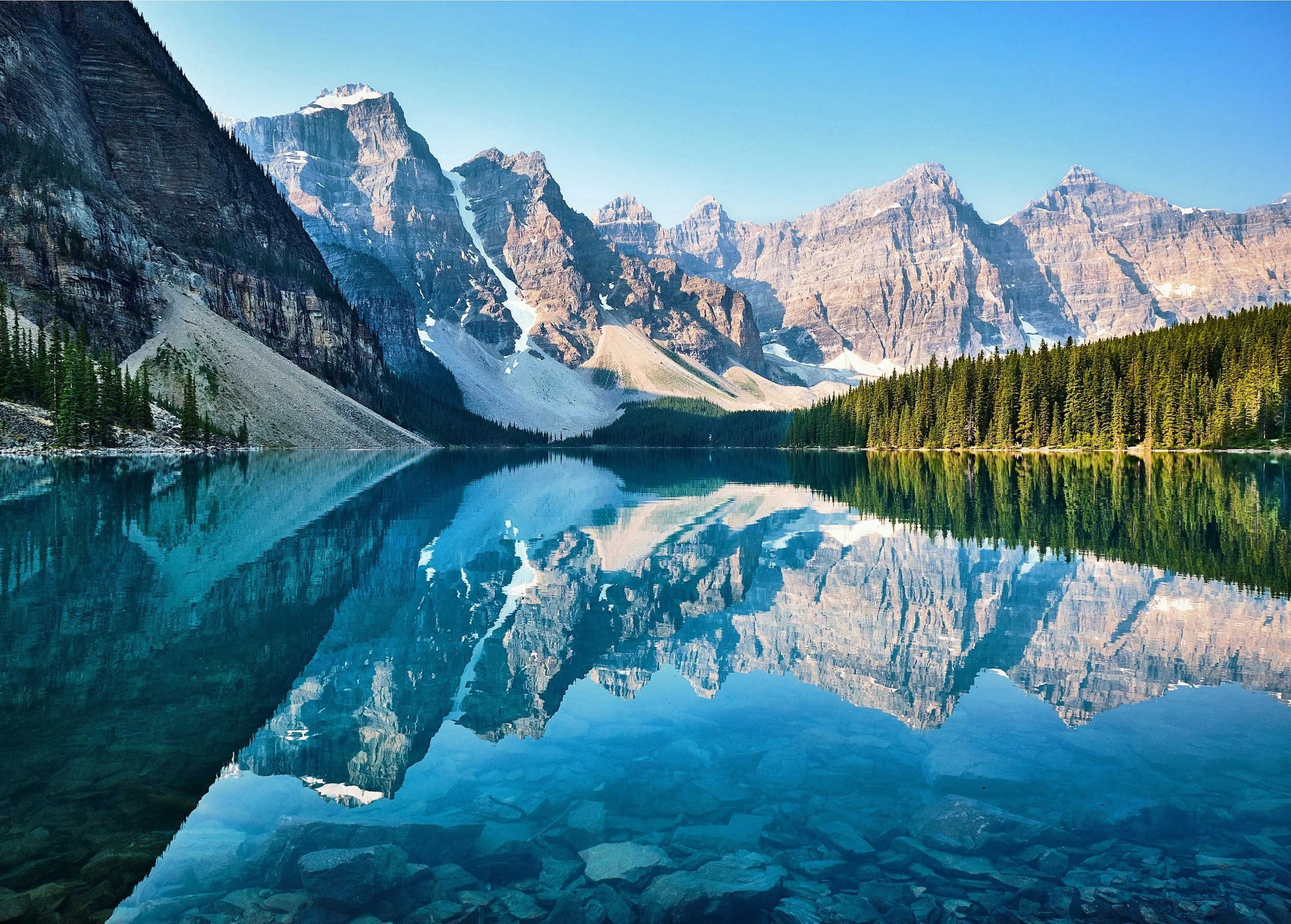Lac de montagne avec eaux calmes reflétant les sommets enneigés, forêt de conifères à la rive et montagnes rocheuses avec peu de neige sous un ciel clair