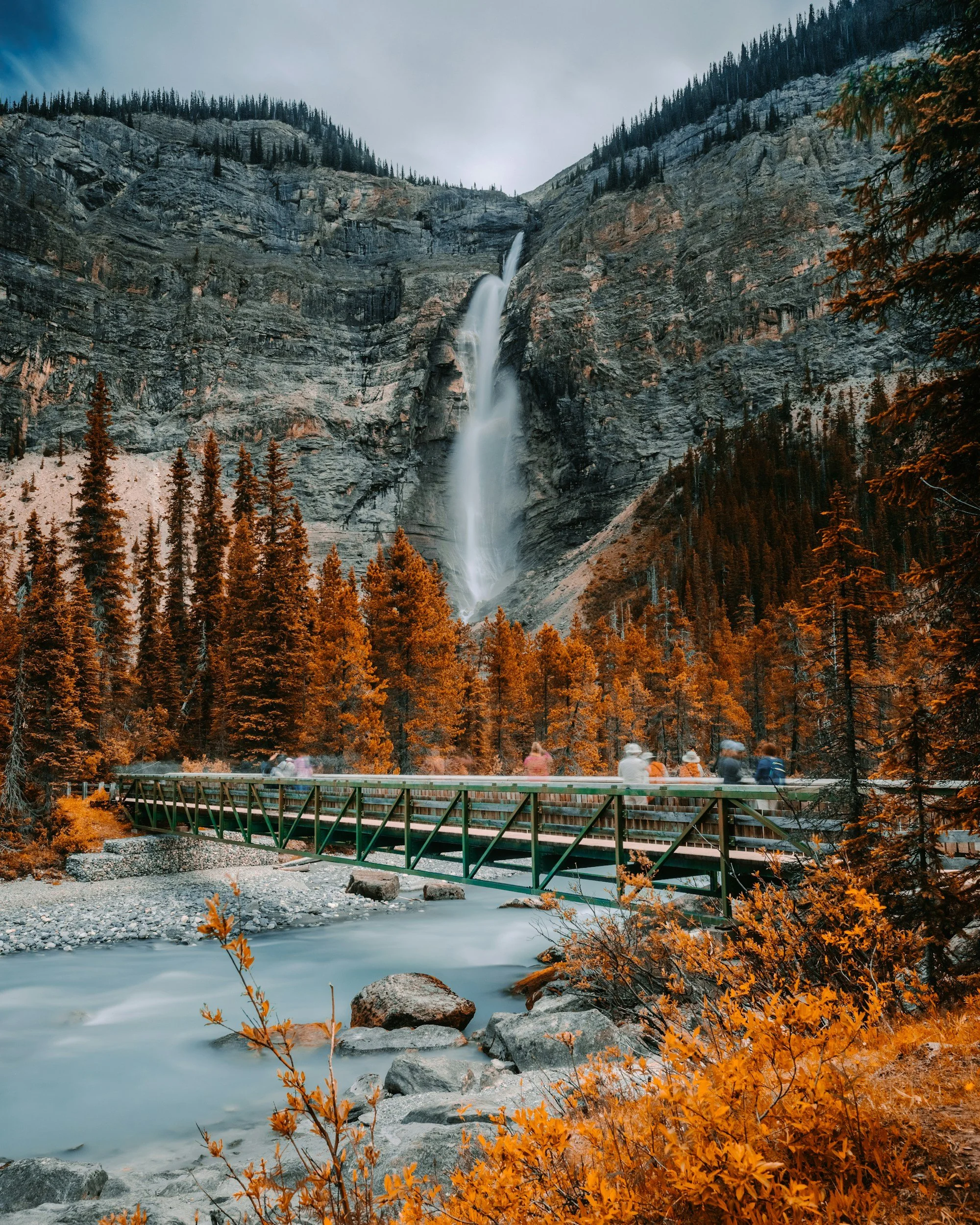 Cascade au cœur d’une forêt automnale avec un pont en bois traversant une rivière. Des montagnes et une chute d’eau en arrière-plan.