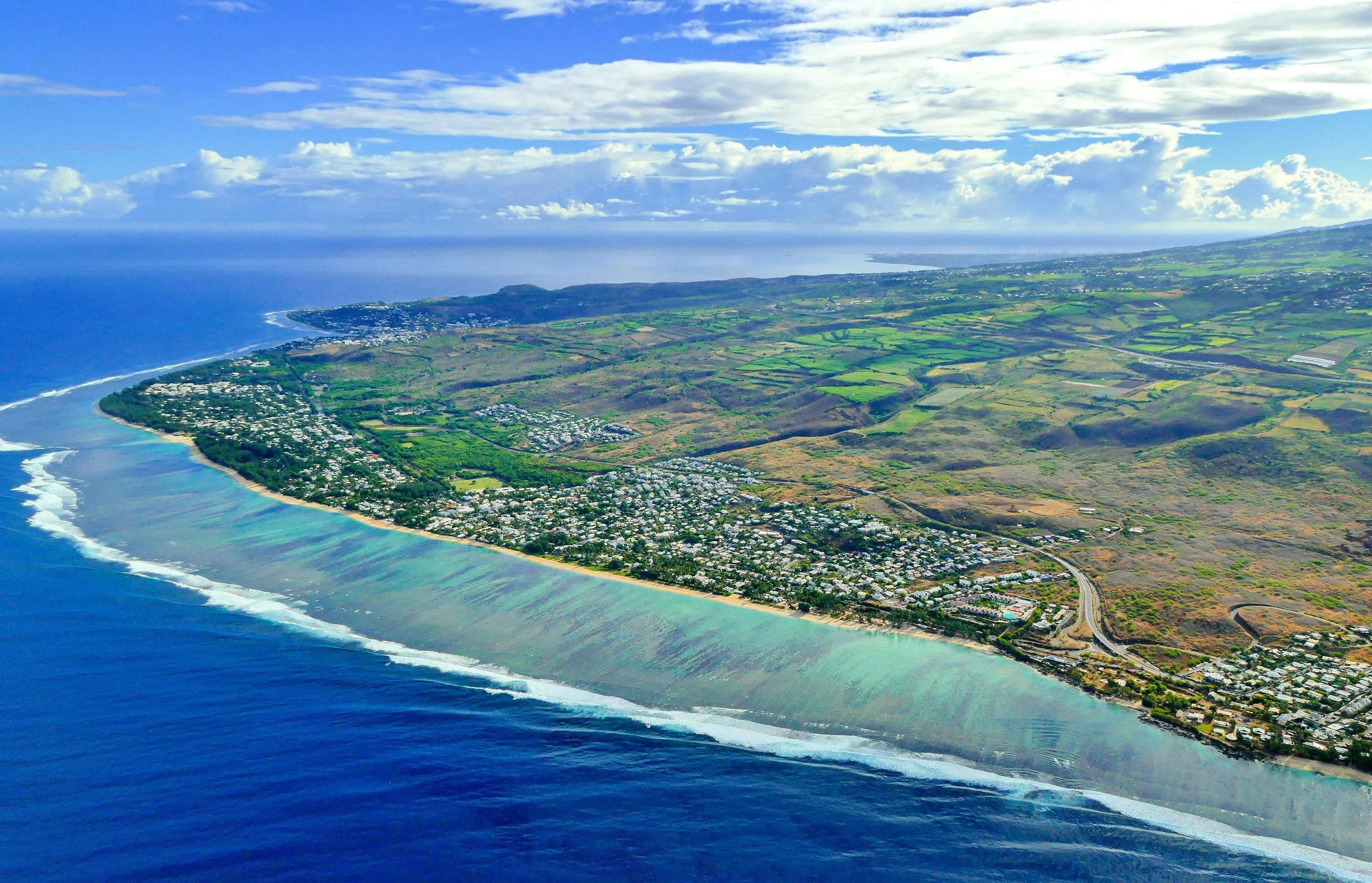 Vue aérienne d'une côte avec plage, l'océan, terrain verdoyant, zones résidentielles et routes dans un paysage insulaire.