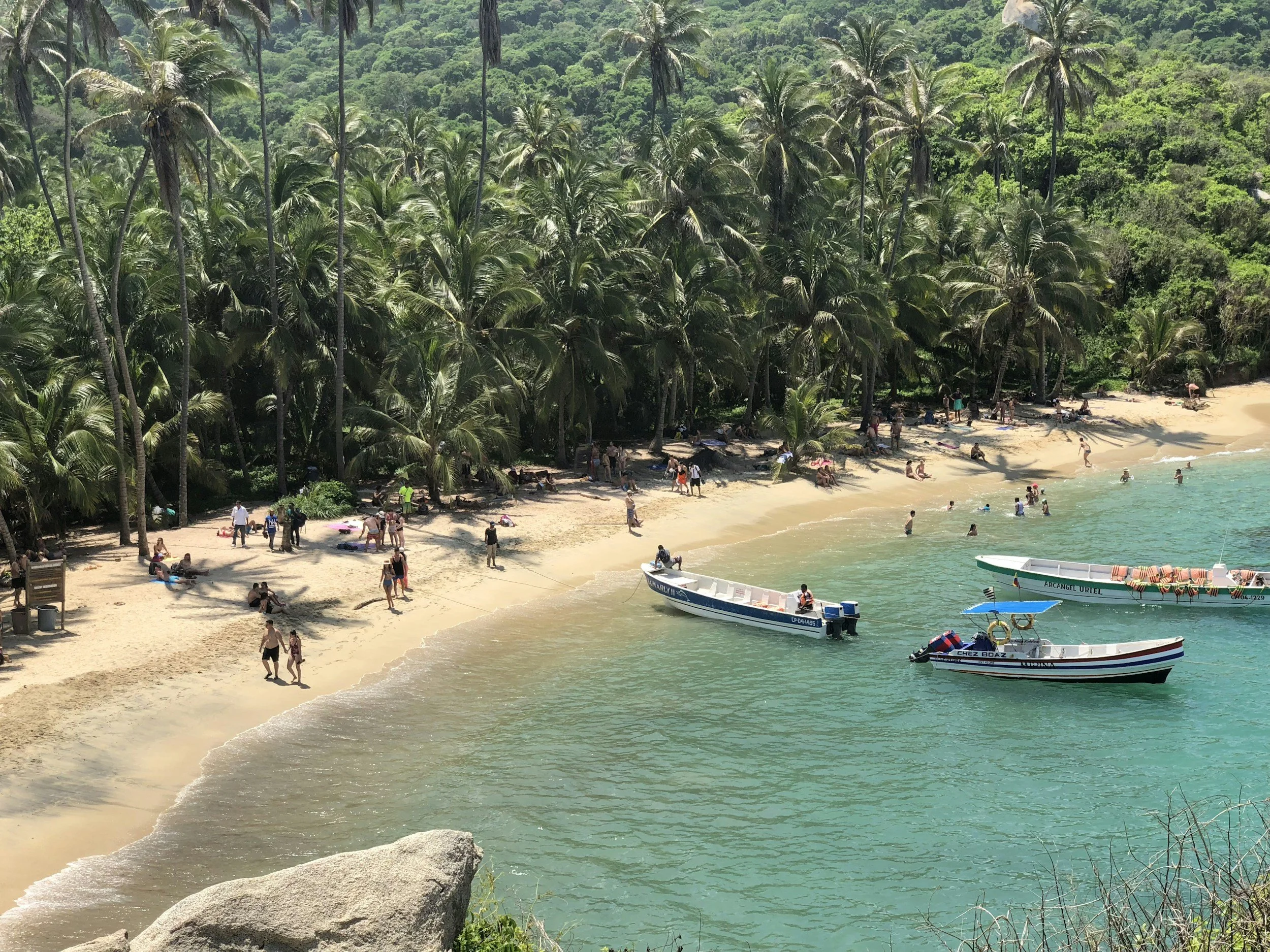 Plage tropicale avec des palmiers, des personnes qui se détendent sur le sable, des bateaux flottant dans la mer claire et une forêt dense à l'arrière-plan.