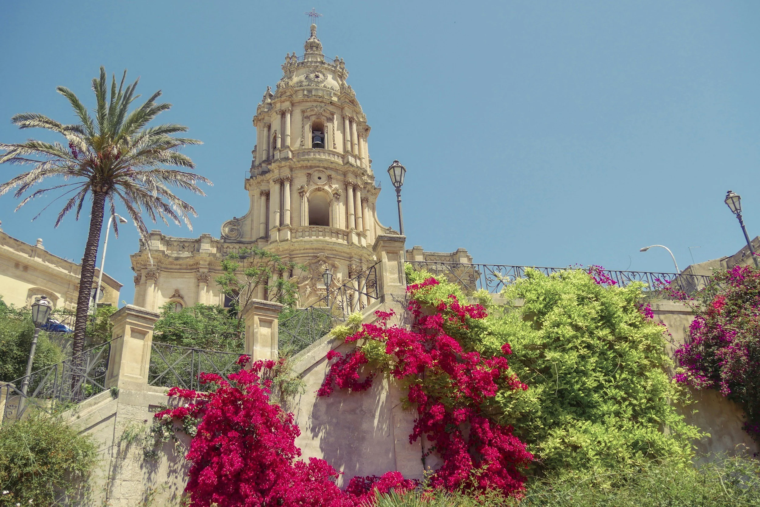 Une église baroque au sommet d'une colline, entourée de palmiers et de fleurs colorées, sous un ciel bleu clair.