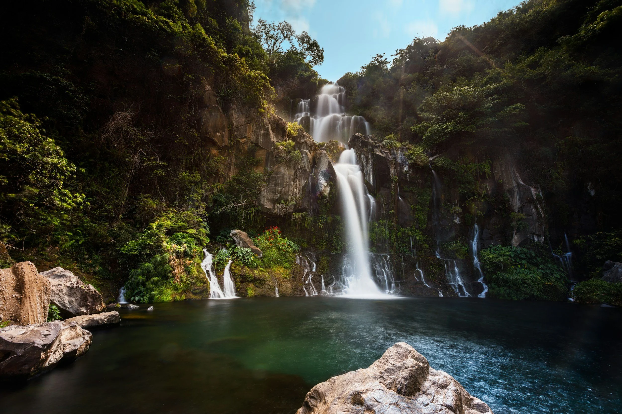 Une cascade dans une forêt luxuriante avec de l'eau qui tombe dans une piscine naturelle, sous un ciel bleu ensoleillé.