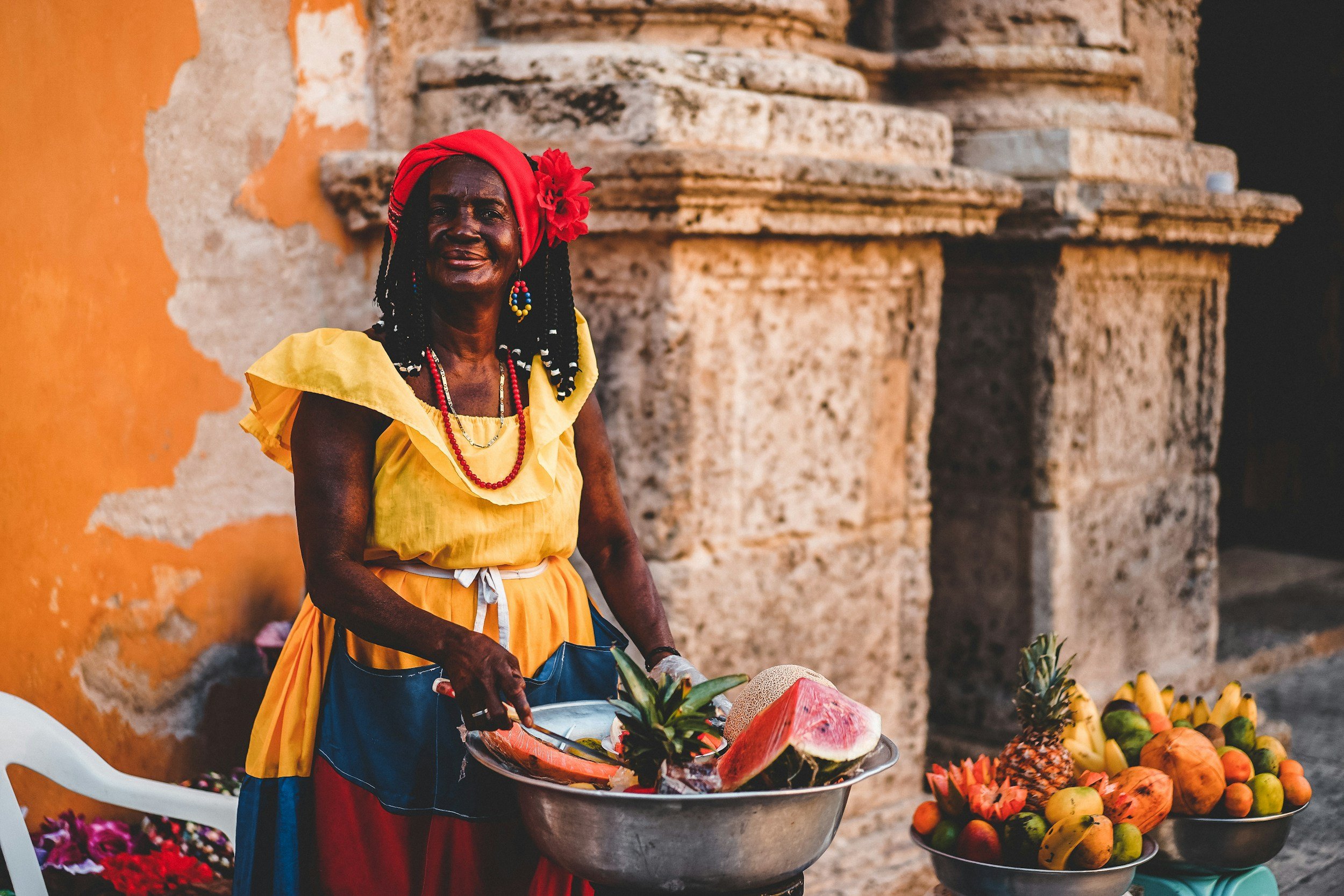 Une femme vendant des fruits et légumes colorés dans un marché, portant des vêtements traditionnels, avec un mur en pierre en arrière-plan.