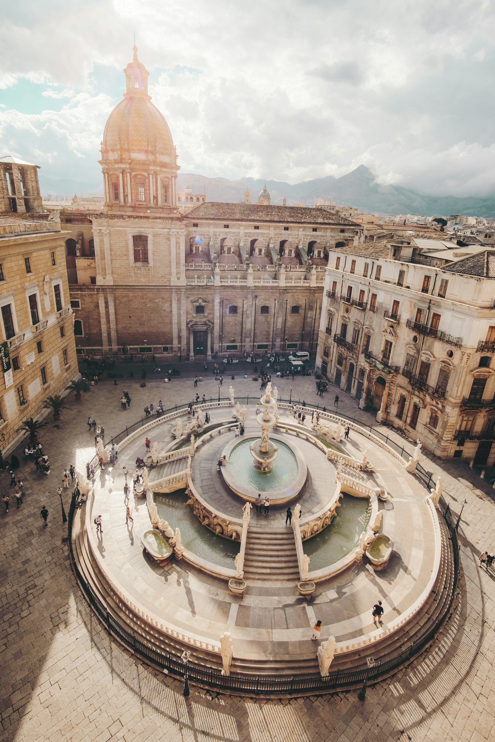 Une fontaine décorative ornée de statues en pierre dans une place urbaine européenne, avec un grand bâtiment historique à dôme en arrière-plan et des montagnes au loin, sous un ciel partiellement nuageux.