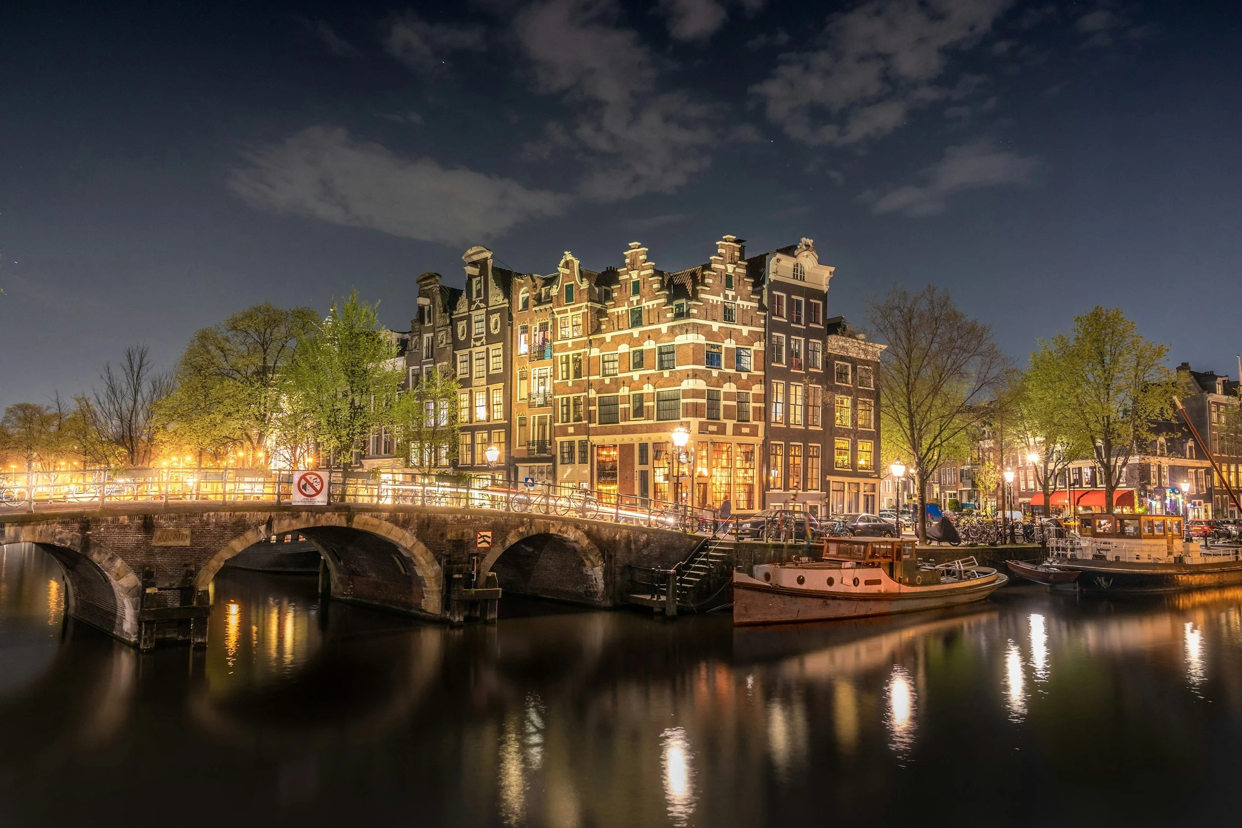 Une vue nocturne d'une ville avec des bâtiments traditionnels le long d'une rivière, des lampadaires éclairant la scène, des arbres verts, et des bateaux amarrés sur l'eau.