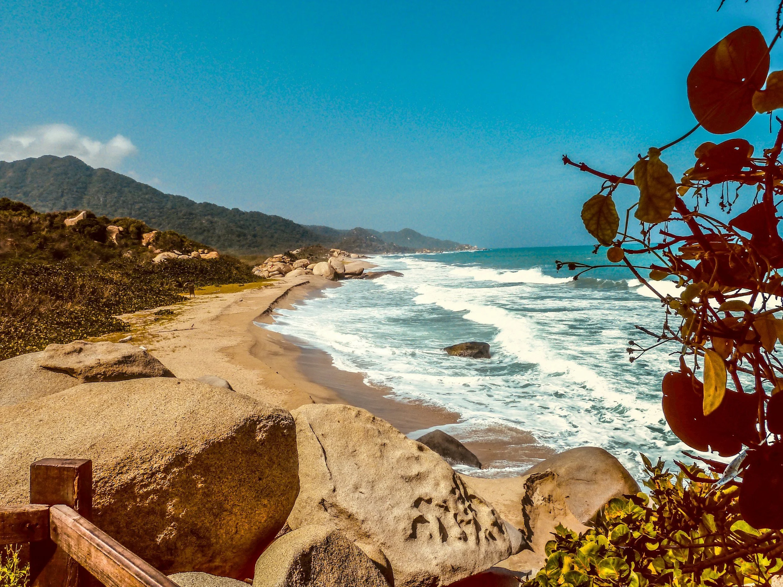 Plage de sable avec des rochers, végétation dense à gauche, montagnes en arrière-plan, mer calme avec vagues, ciel bleu clair.