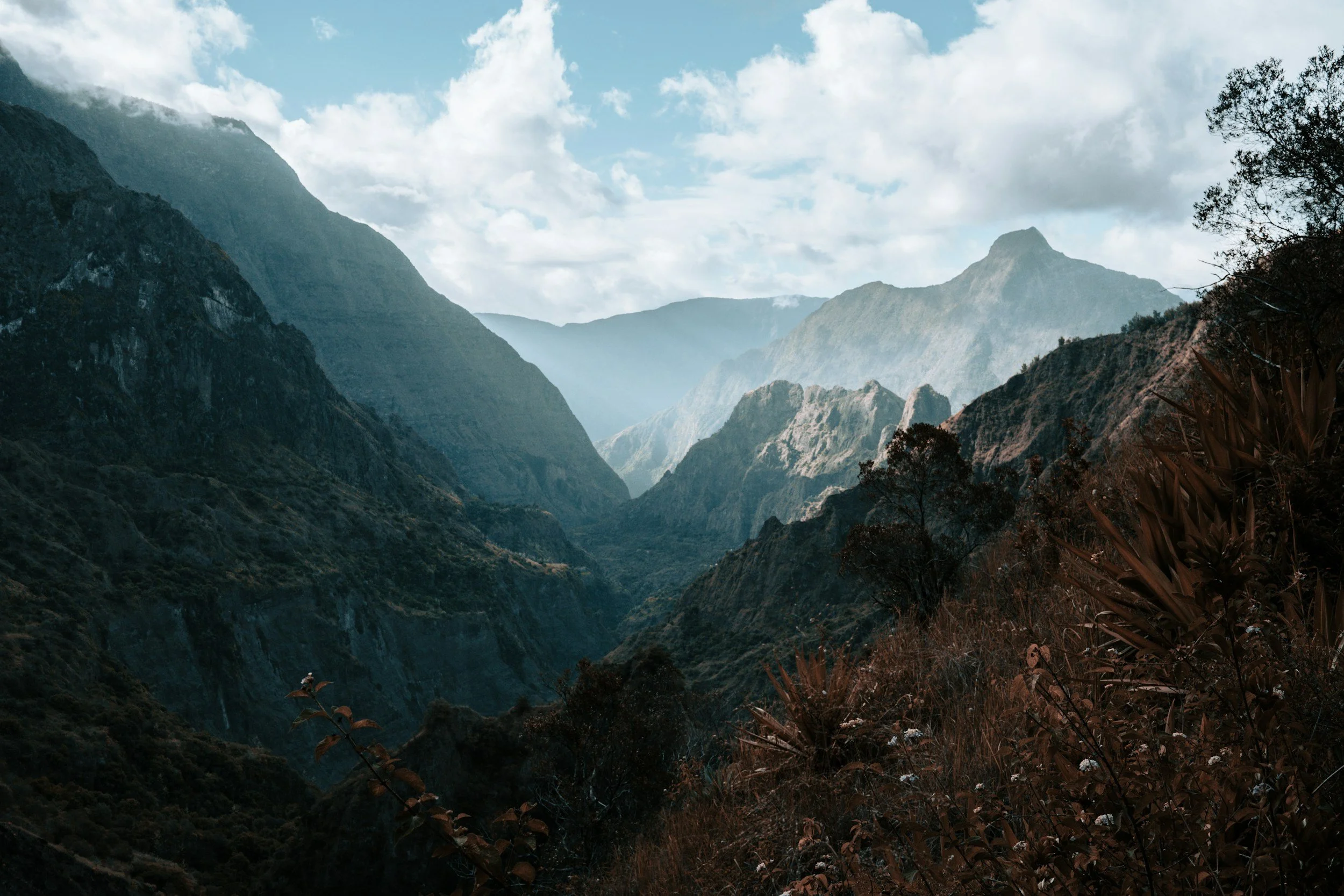 Vue d'une vallée de montagne avec des sommets enneigés en arrière-plan, des montagnes escarpées, un ciel partiellement nuageux, et de la végétation dans la partie inférieure de l'image.