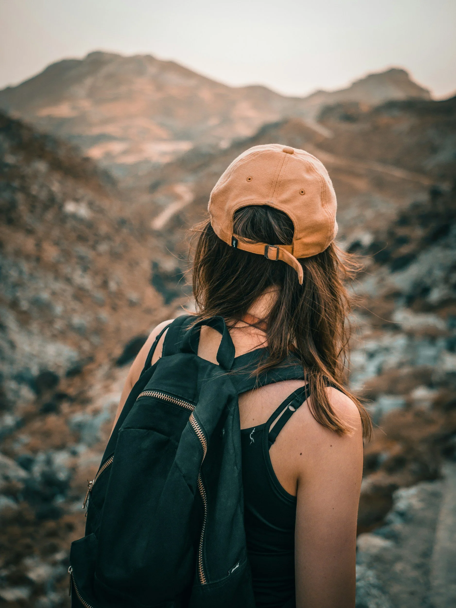 Une femme avec un sac à dos, portant une casquette beige, vue de dos, dans un paysage montagneux aride.