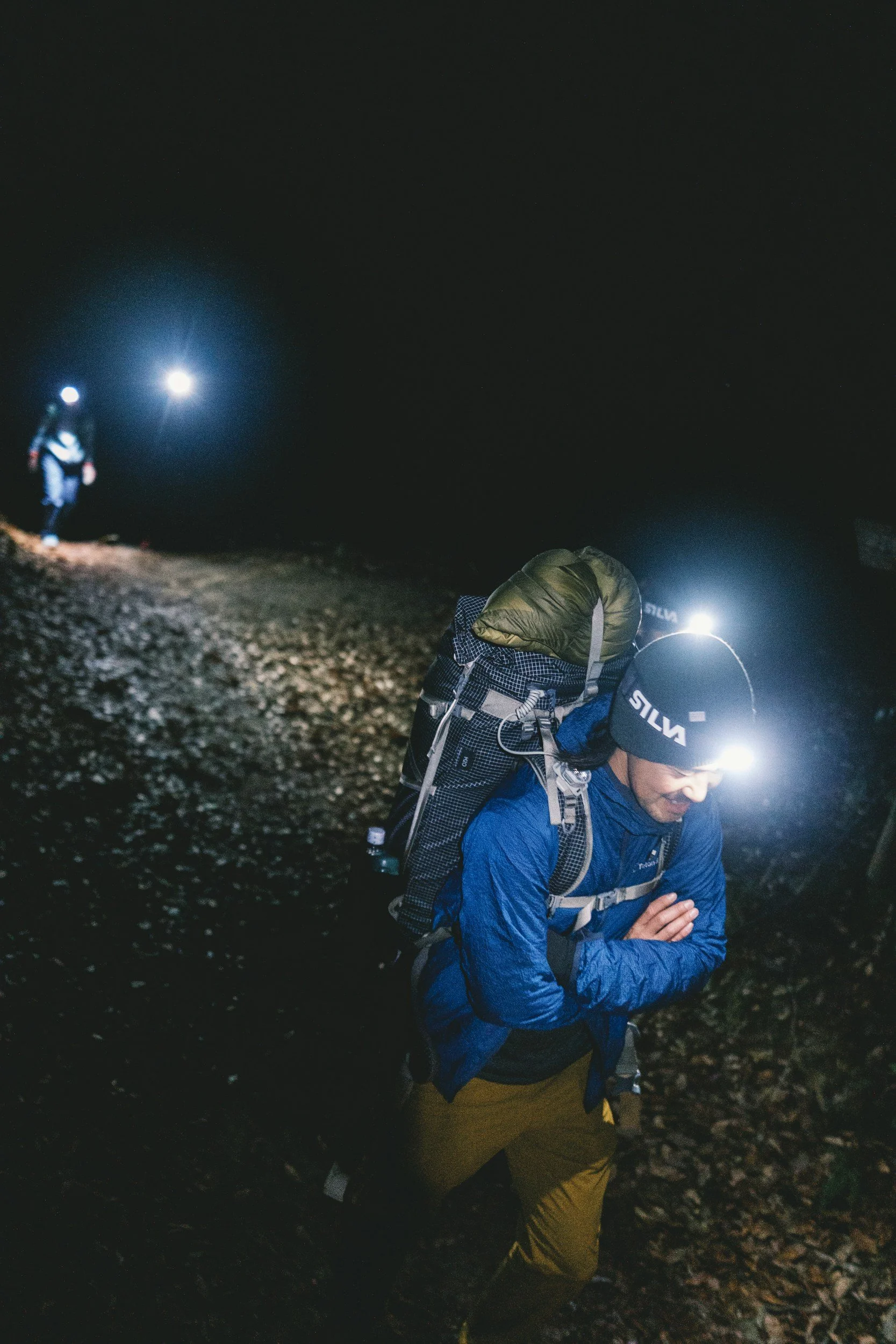 Un homme avec une tête lampée, portant un sac à dos, marche dans un environnement nocturne avec un autre randonneur en arrière-plan.