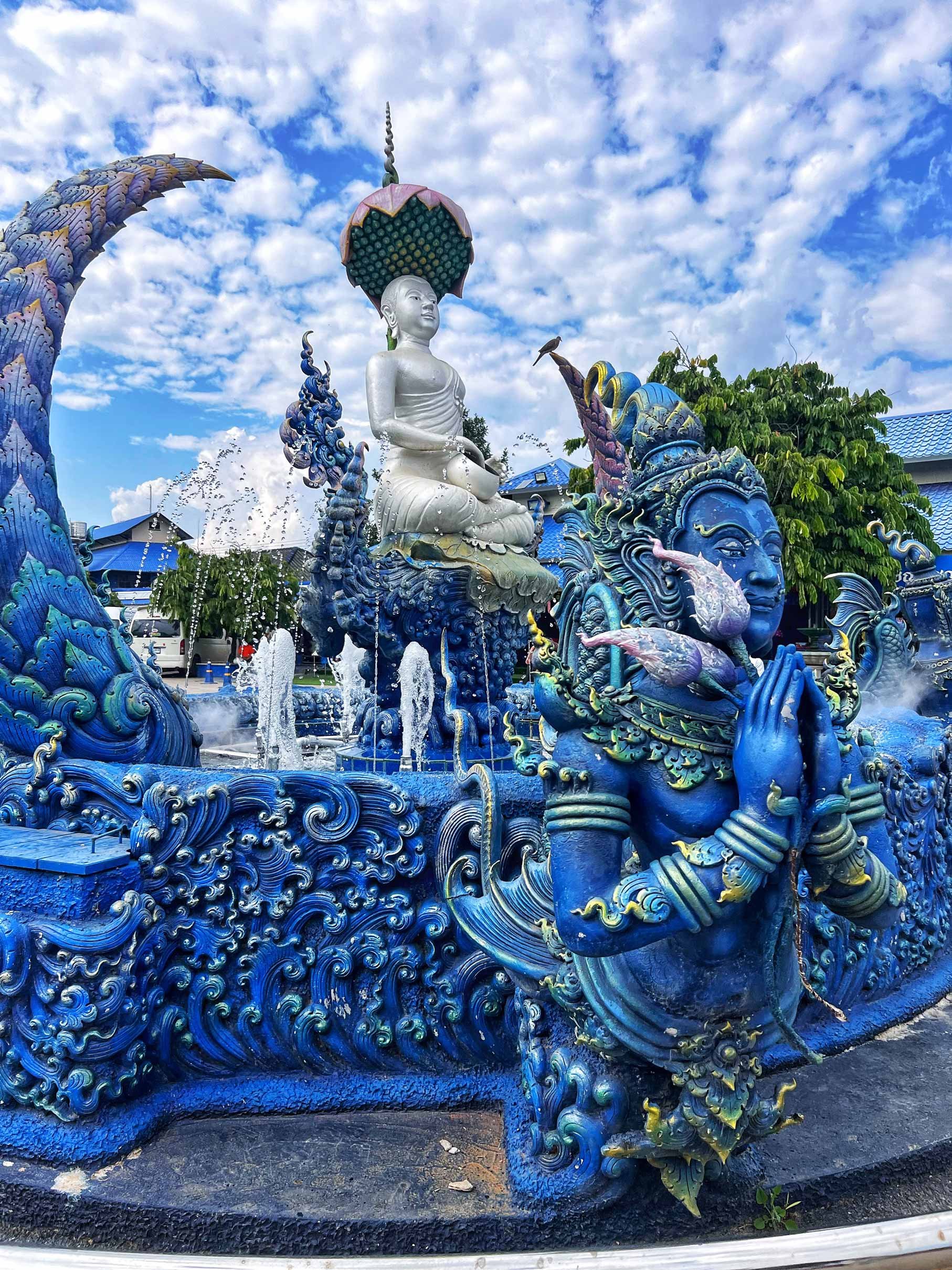 Une fontaine ornée de sculptures colorées représentant des figures mythologiques et des motifs aquatiques sous un ciel bleu avec quelques nuages.