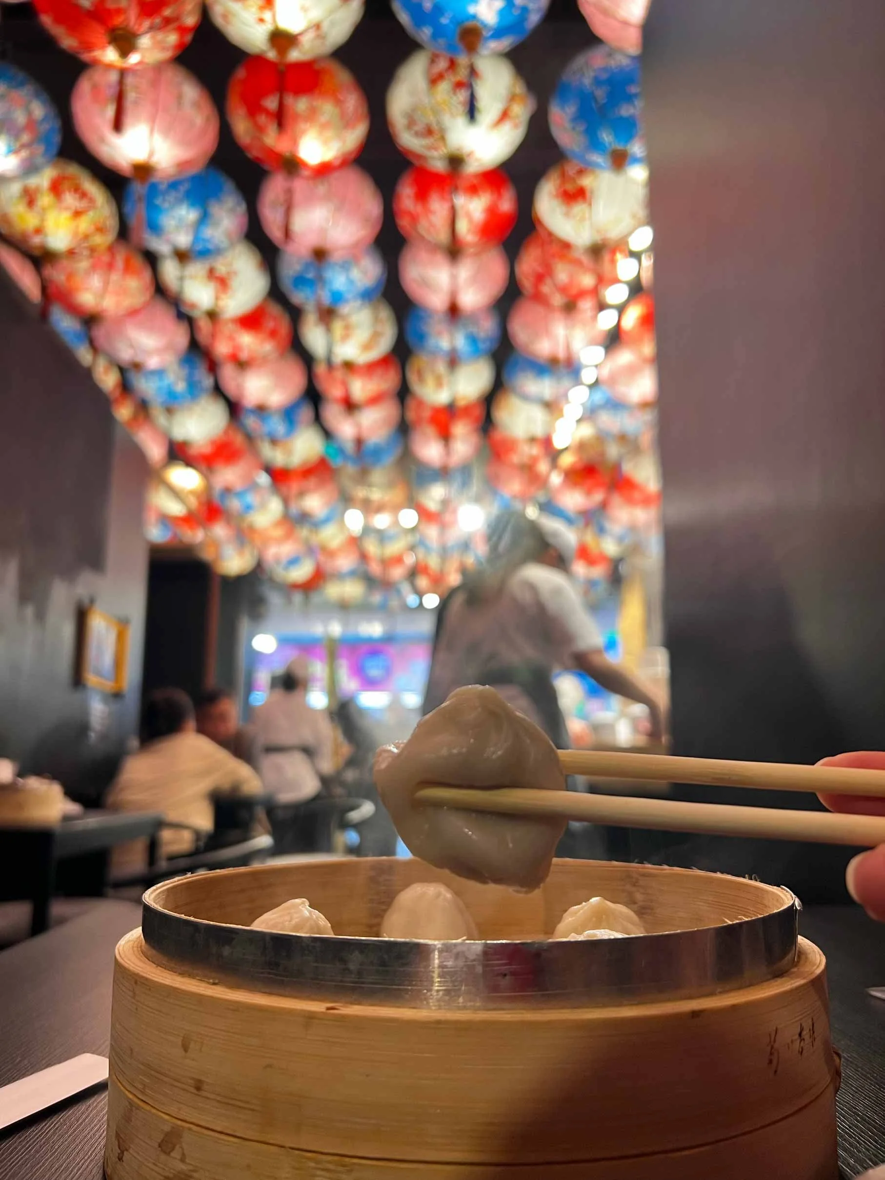 Dans un restaurant, on voit un panier en bambou contenant des raviolis à vapeur, une personne utilise des baguettes pour prendre un ravioli. Le plafond est décoré de lanternes rouges et blanches avec des motifs colorés, et il y a d'autres personnes en arrière-plan.