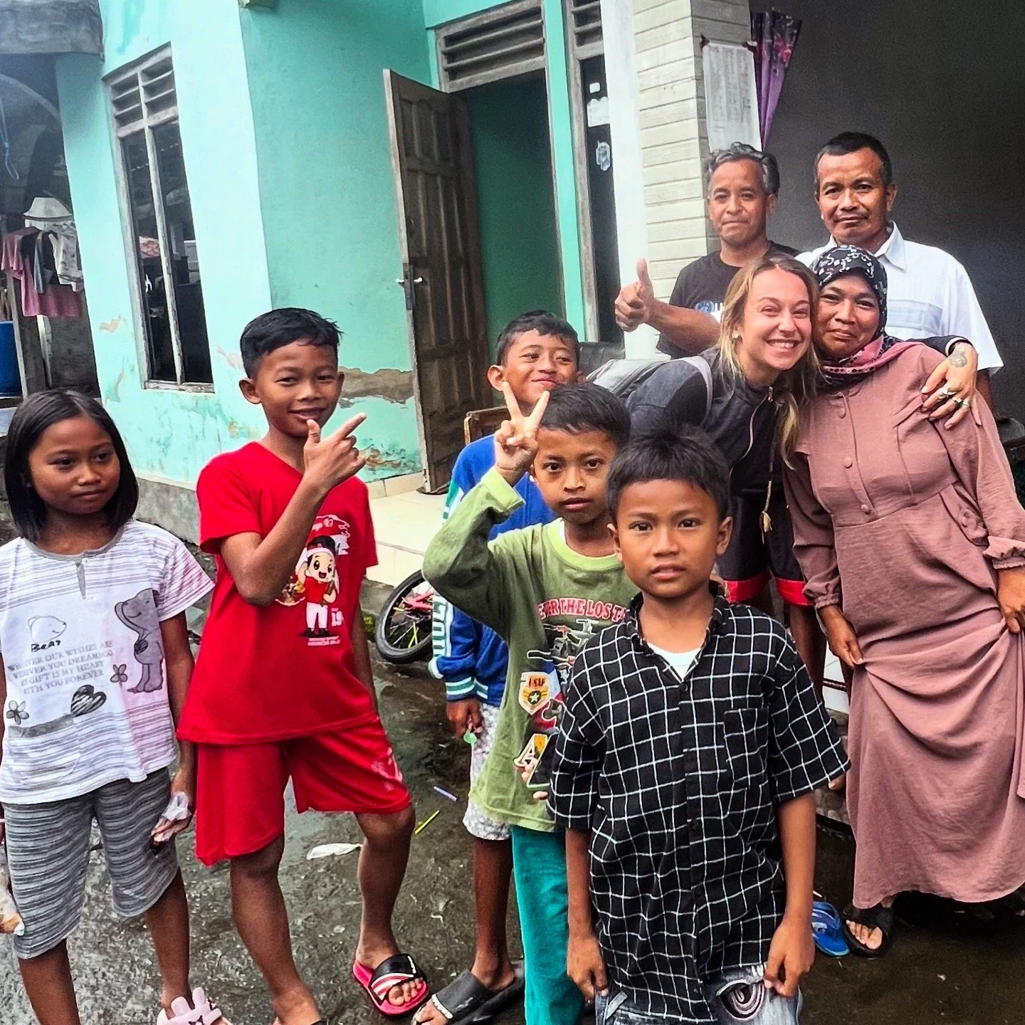 Groupe de personnes, enfants et adultes, se rassemblant devant une maison, certains faisant des signes de paix, en milieu urbain.