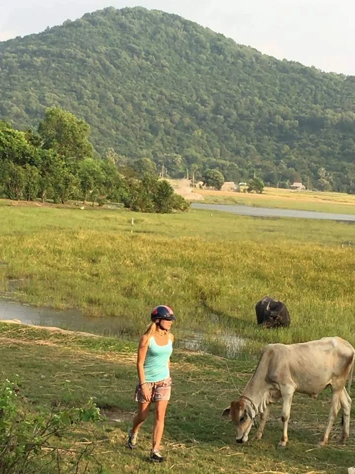 Une femme portant un casque de moto se promène près d'une vache dans un paysage rural avec des montagnes en arrière-plan, de l'herbe verte et un étang.