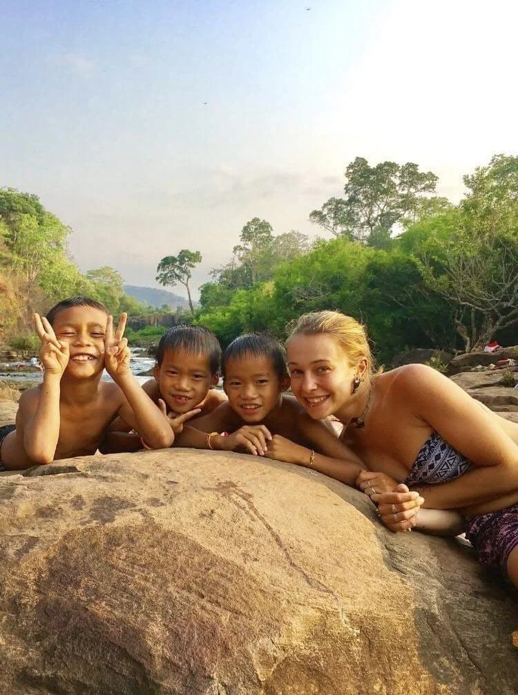 Groupe de quatre personnes (trois enfants et une femme) allongées sur une roche près d'une rivière, entourées de végétation luxuriante, en milieu naturel