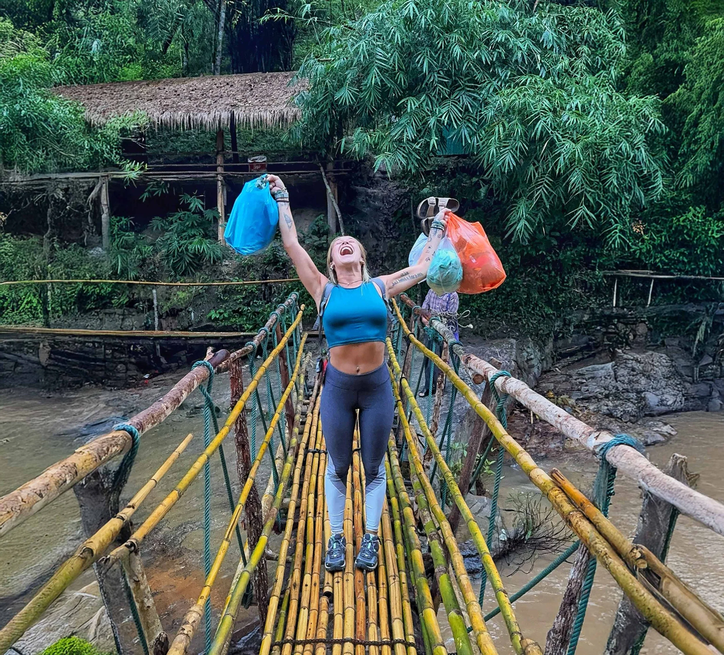 Charlotte souriante, en tenue de sport, traverse un pont suspendu en forêt avec des sacs à main colorés dans chaque main, entourée d'une végétation dense.