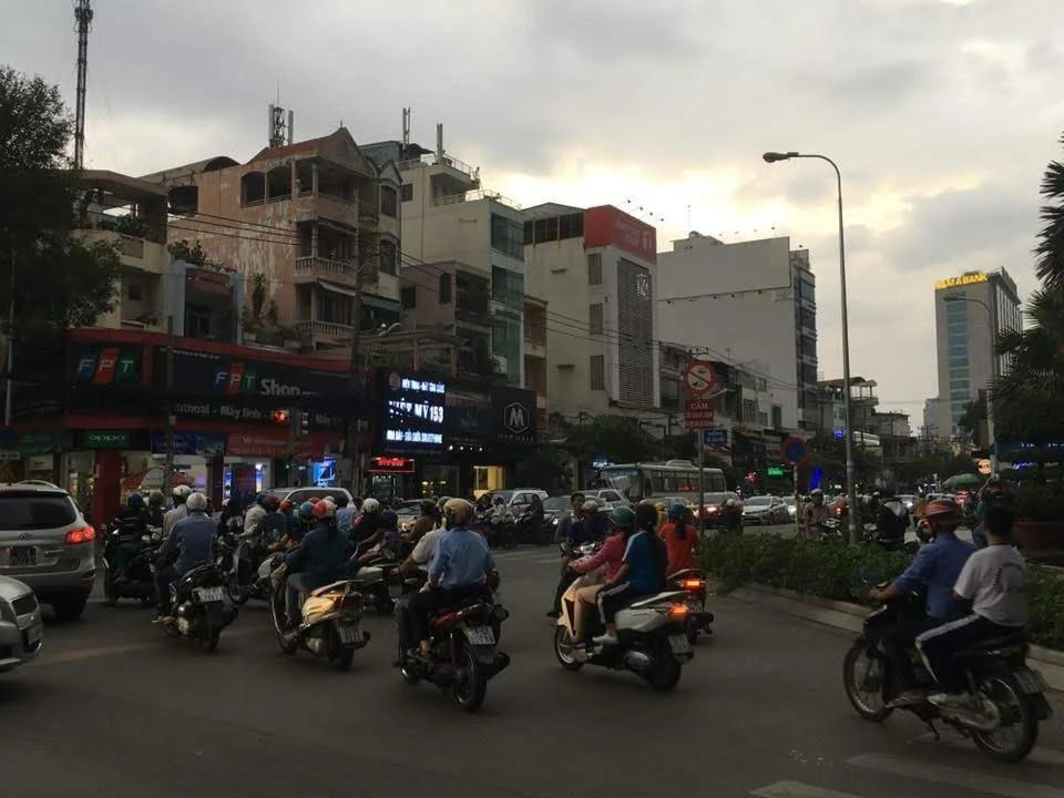 Une scène urbaine avec beaucoup de motos et quelques voitures en circulation, devant des bâtiments commerciaux et résidentiels, sous un ciel nuageux en fin d'après-midi ou début de soirée.