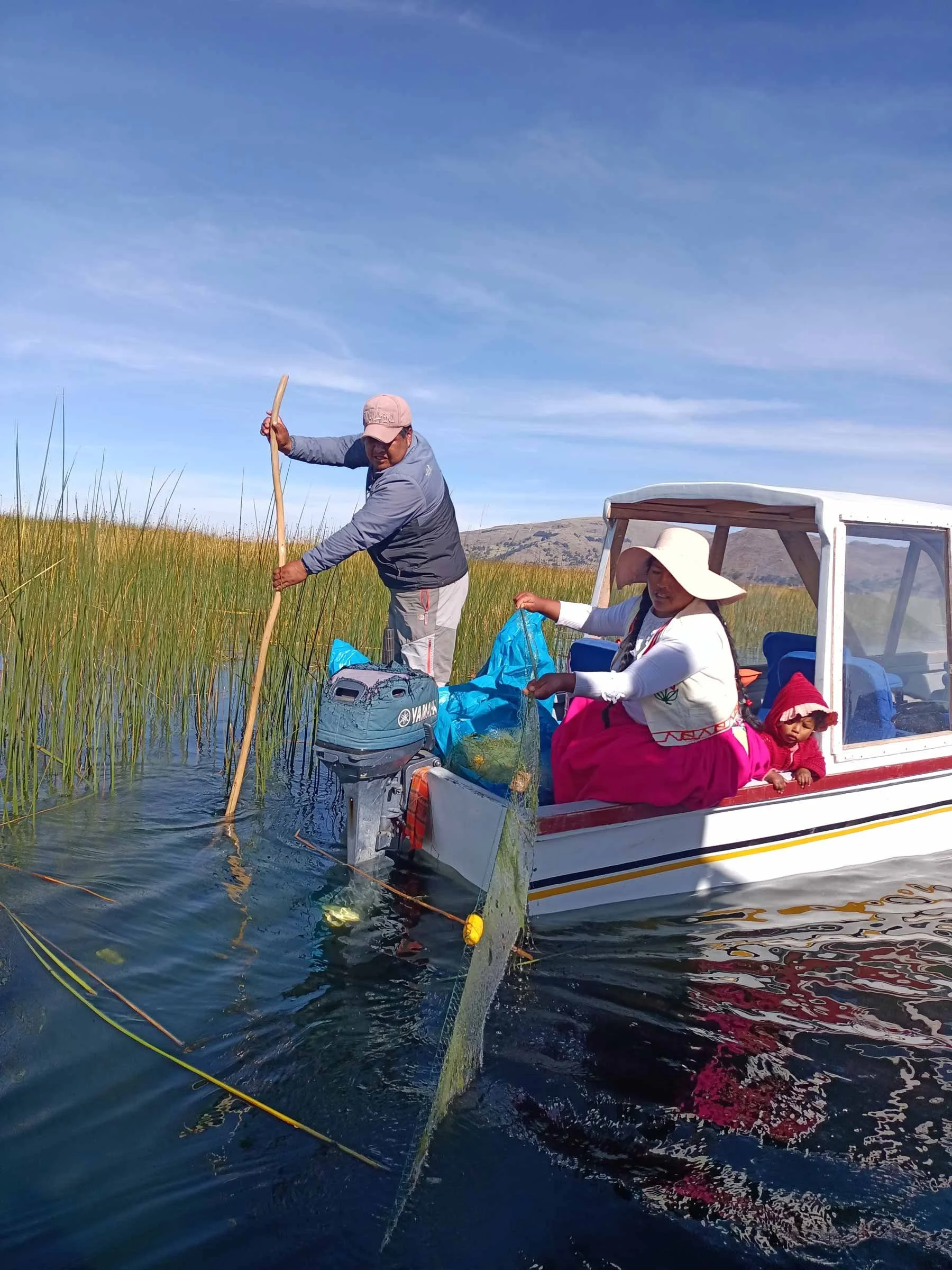 Trois personnes, deux adultes et une enfant, en train de pêcher à bord d'une petite embarcation dans un lac ou une rivière, avec des plantes aquatiques en bordure et un paysage montagneux en arrière-plan.