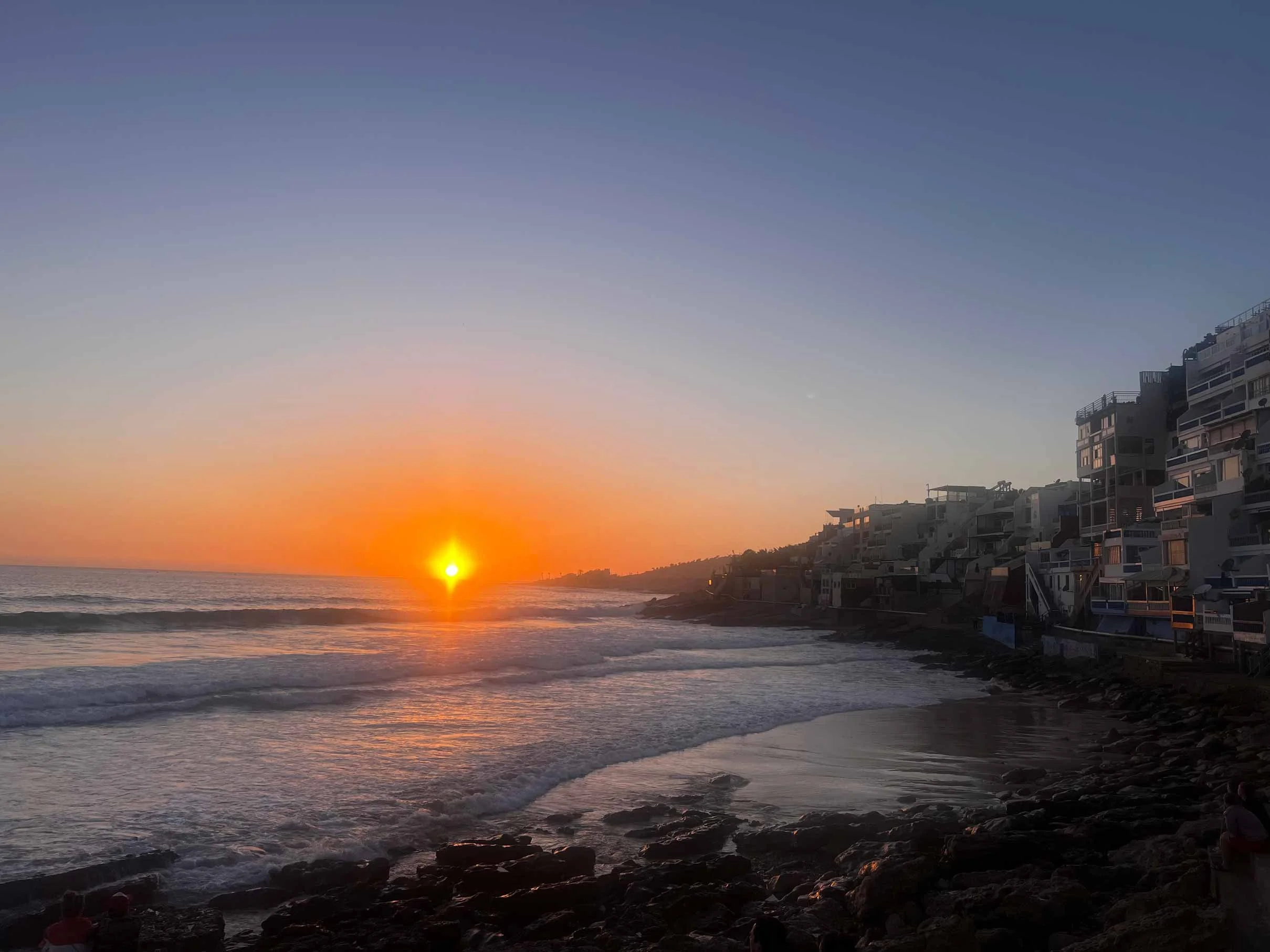 Coucher de soleil sur la plage avec des bâtiments résidentiels en arrière-plan, lune visible dans le ciel, vagues calmes et rochers au premier plan.