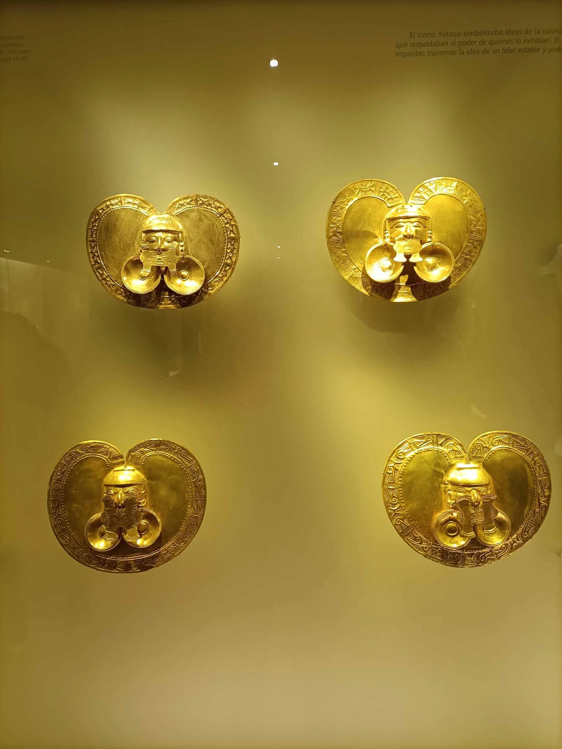 Quatre bijoux dorés avec un design en forme de cœur, suspendus contre un fond jaune, exposés dans une vitrine de musée.