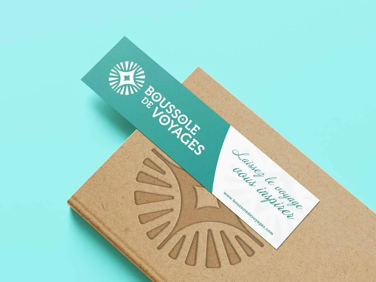 Carton en kraft et étiquette bleue avec logo de boussole de voyage, texte en français "Laissez le voyage nous imprégier" et site web "boussoledevoyages.com" sur un fond bleu clair.