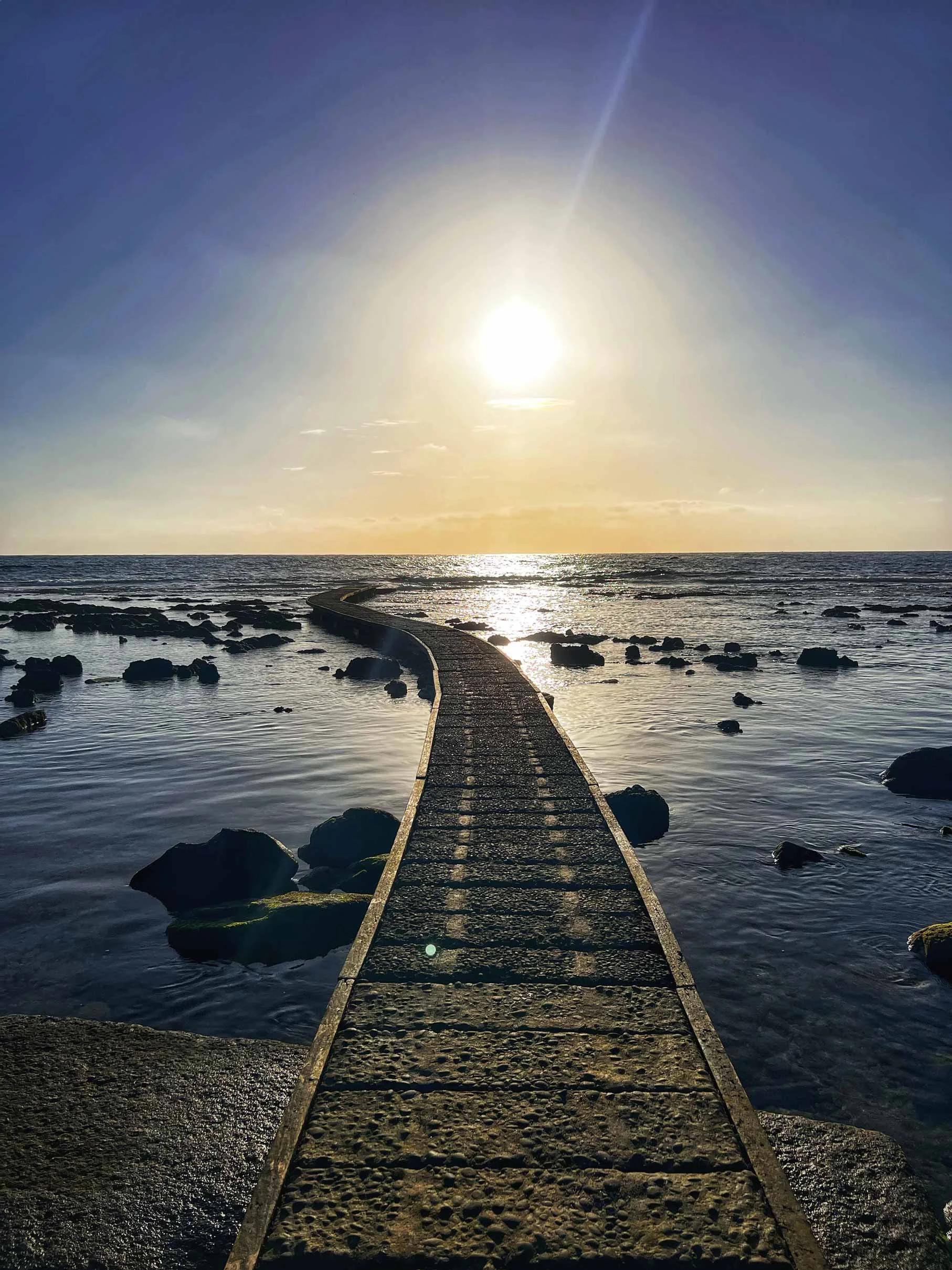 Passerelle en pierre menant à la mer avec des rochers autour, soleil couchant ou levant, reflet dans l'eau, ciel clair.