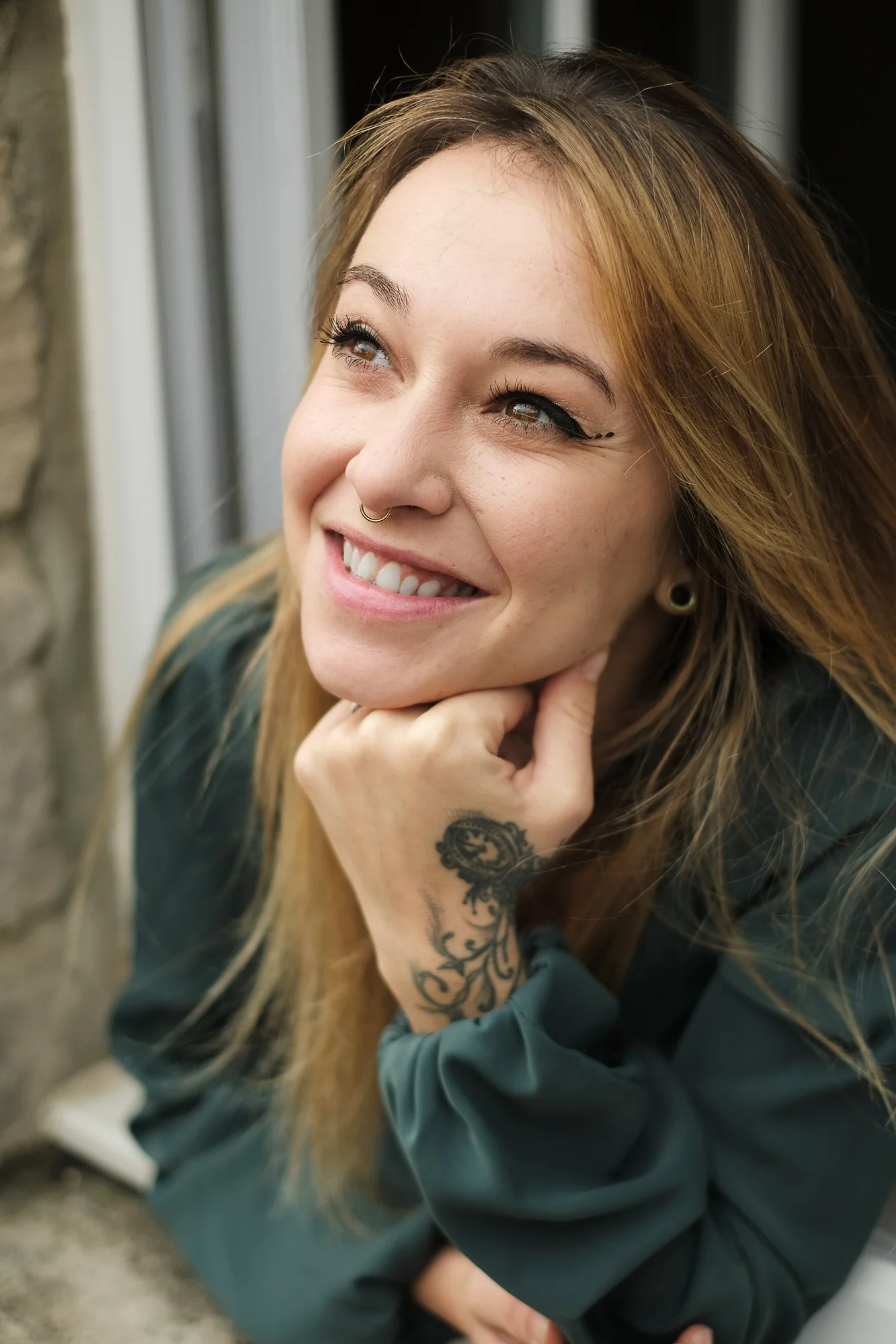 Jeune femme souriante avec tatouage, regardant vers le haut, posant avec la main sous le menton, cheveux longs et roux, portant un vêtement sombre.