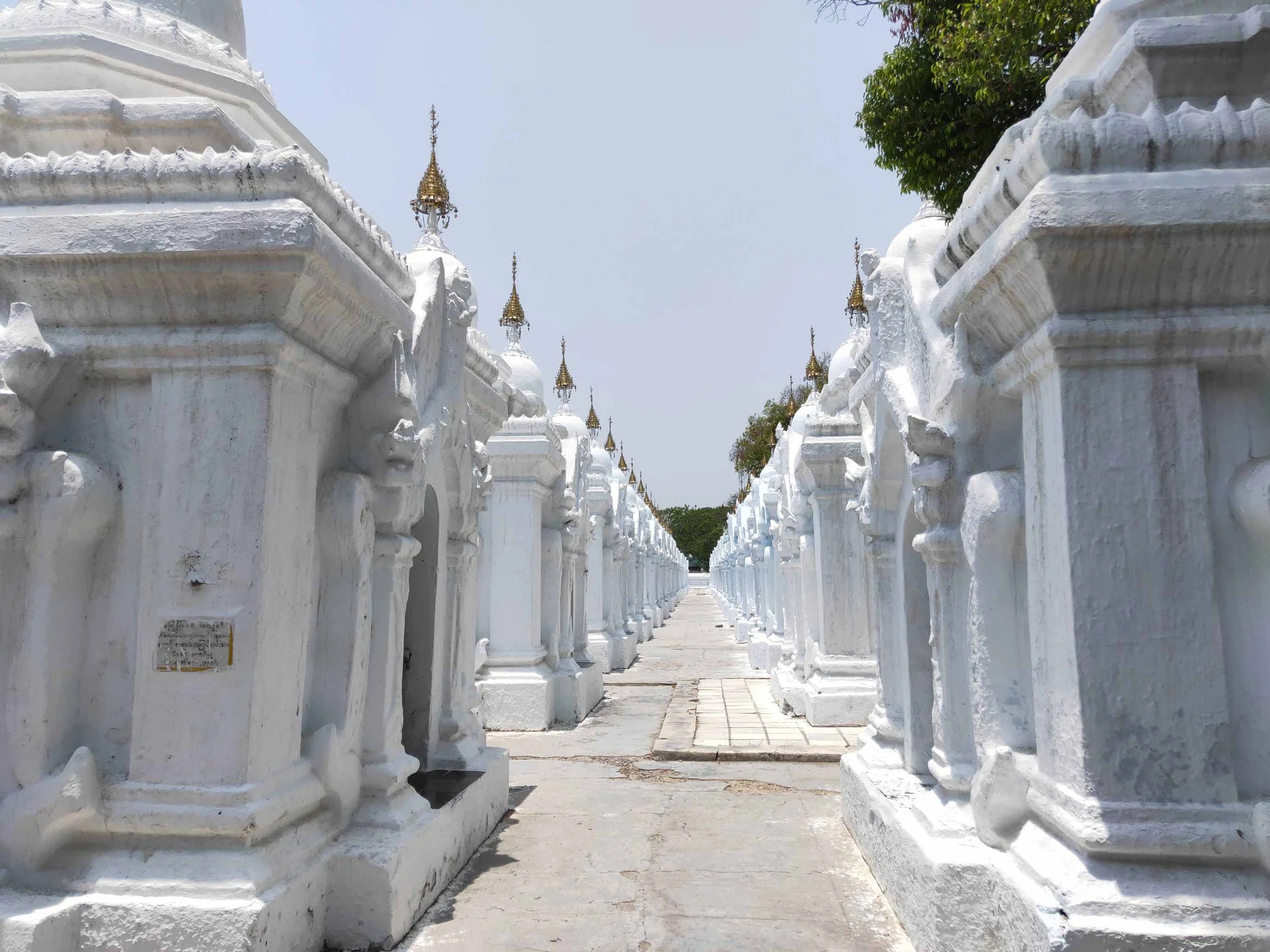 Allée de stupas blancs ornés de petites épées dorées, sous un ciel clair, dans un site religieux en Thaïlande.