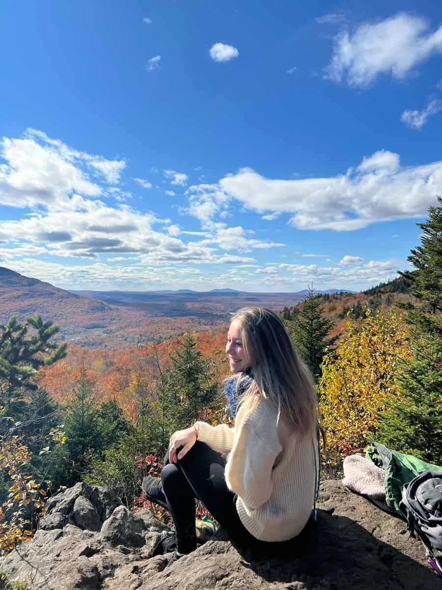 Une femme assise sur un rocher en haut d'une colline, admirant un paysage de montagnes et de forêt aux couleurs automnales, sous un ciel bleu parsemé de nuages.
