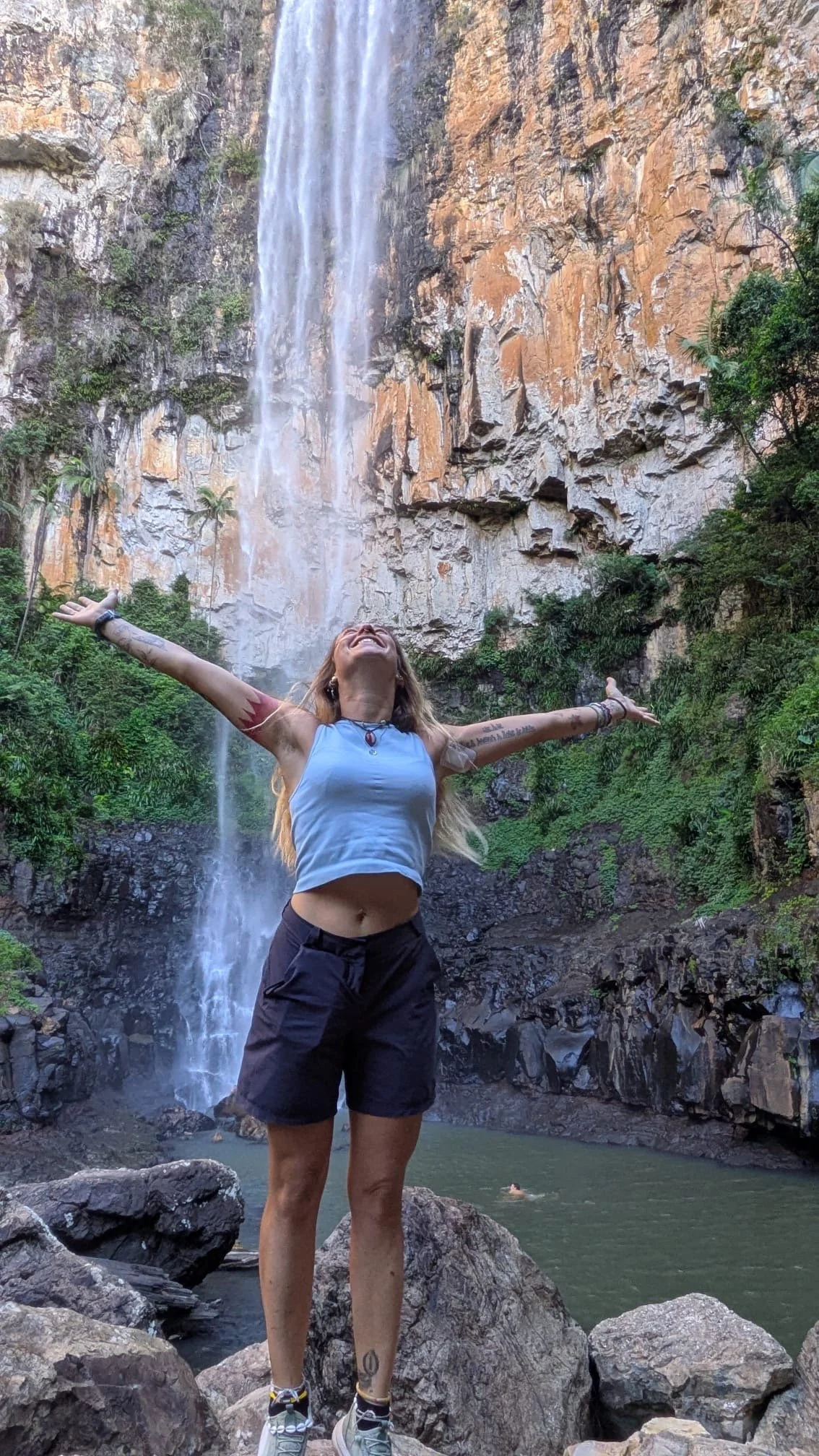 Une femme joyeuse avec les bras ouverts devant une cascade dans une forêt.