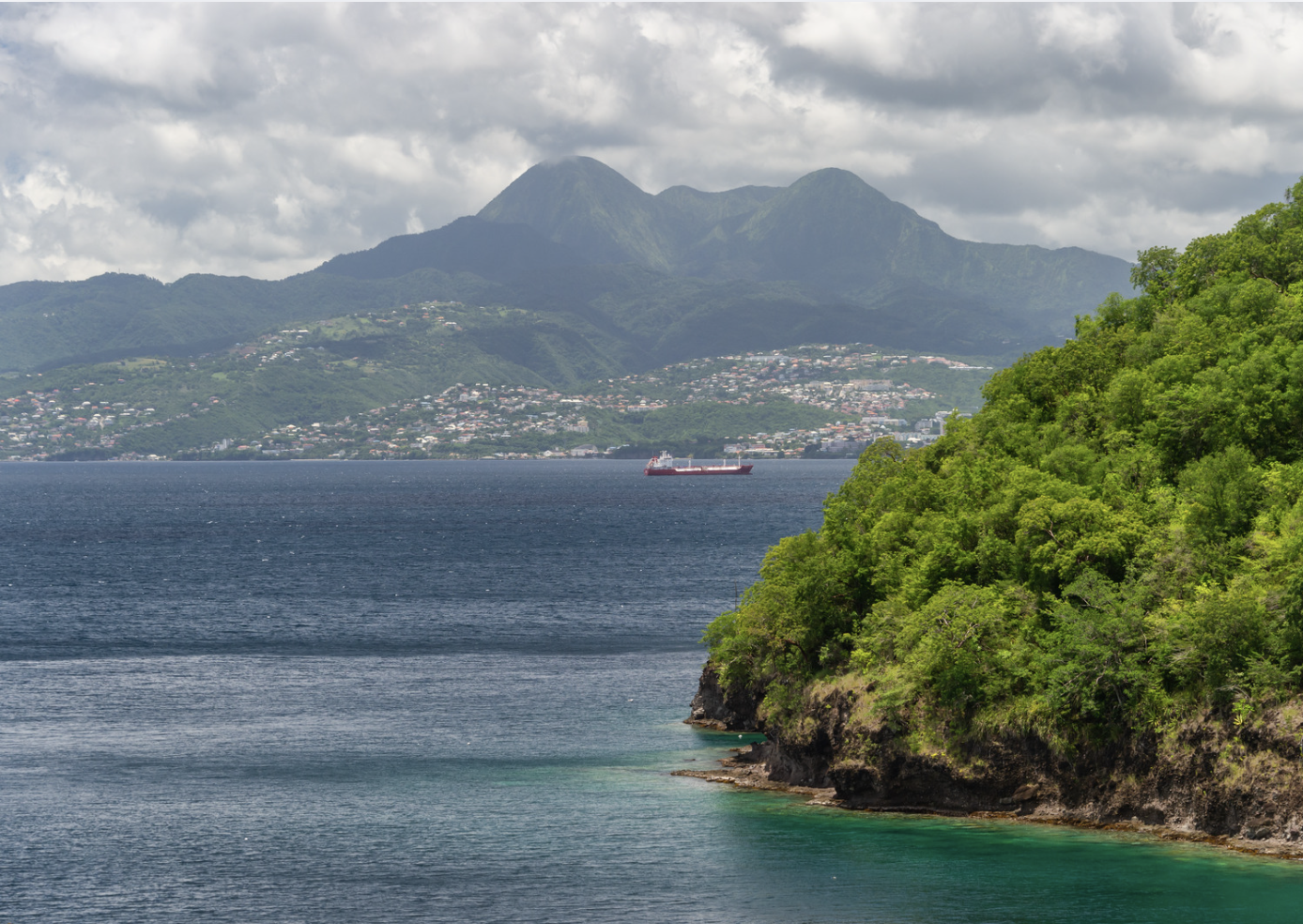 Paysage de mer avec une colline verdoyante au premier plan, une montagne en arrière-plan, un bateau sur la mer, et une ville en contrebas sur la colline.