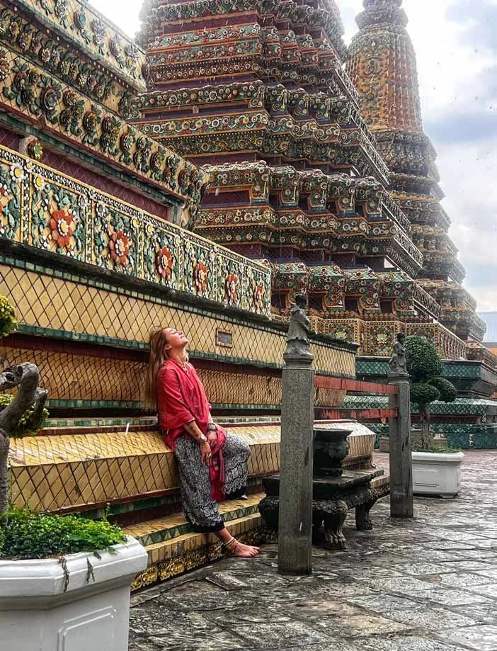 Une femme assise sur un banc décoré à côté d'un temple thaïlandais orné de mosaïques colorées et de sculptures complexes.