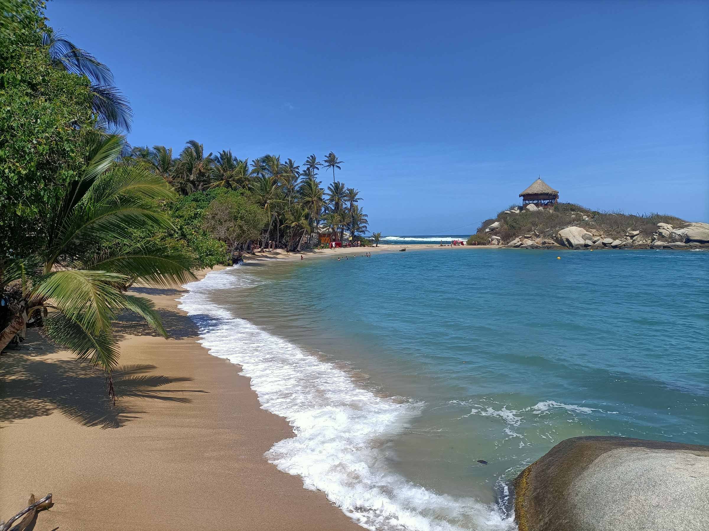 Plage tropicale avec sable doré, palmiers, eau turquoise, et un puit de vue avec hutain en hauteur sur une petite île ou écueil rocheux.